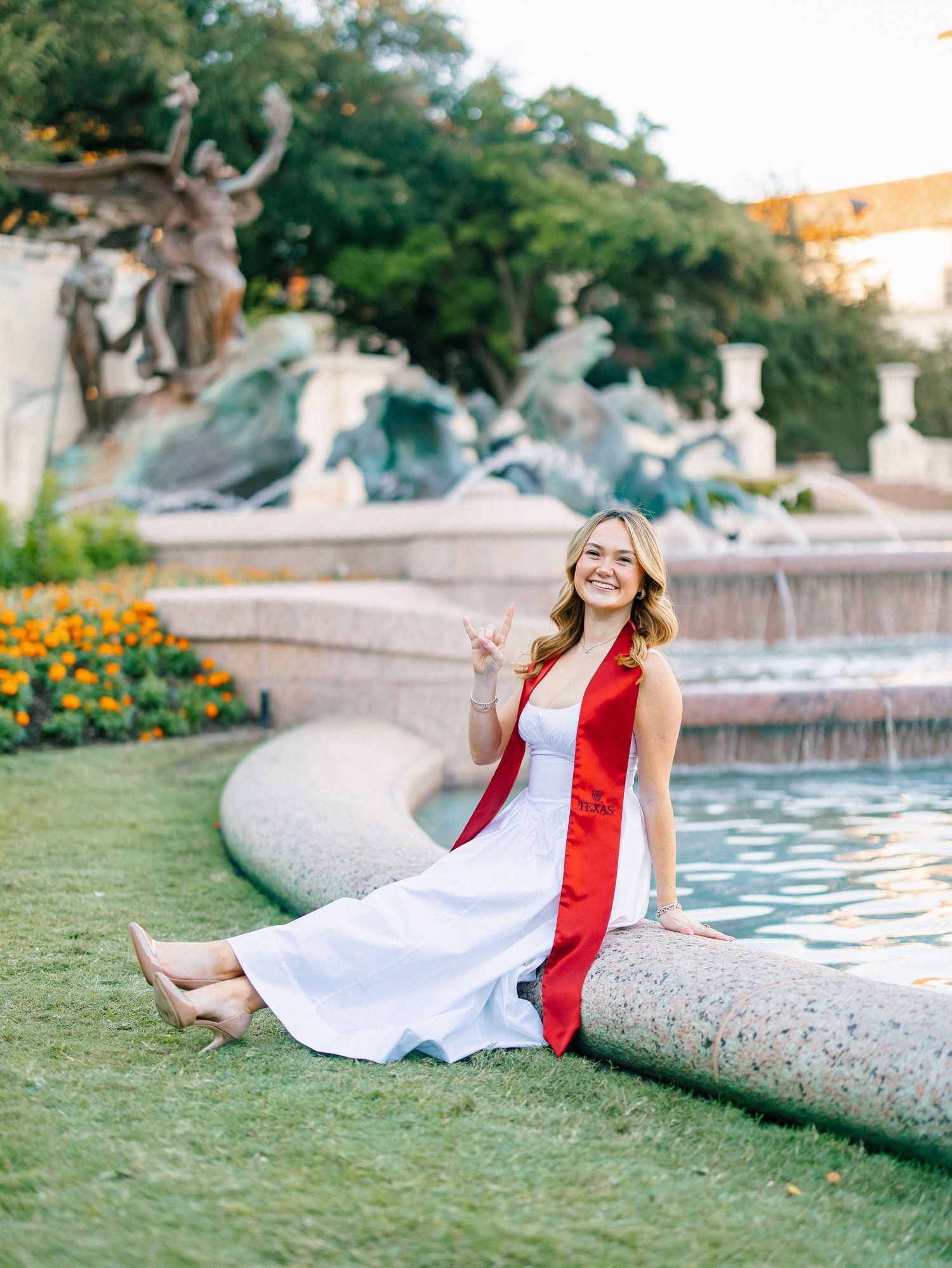 Smiling graduate in white dress and red sash sits by ornate fountain, flashing hand sign on green lawn.