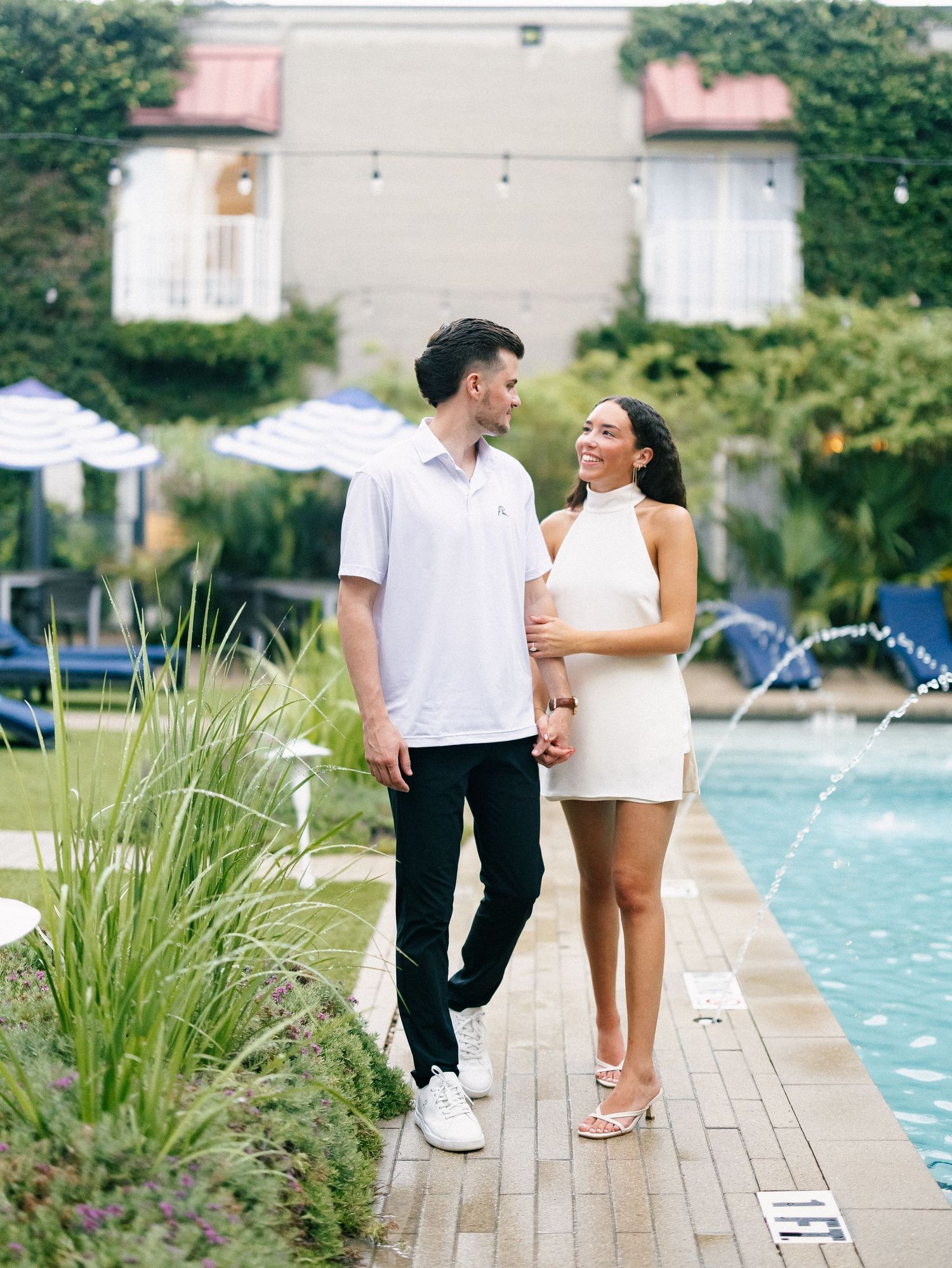 Couple in white outfits walking hand-in-hand beside a pool with fountains and lush greenery.
