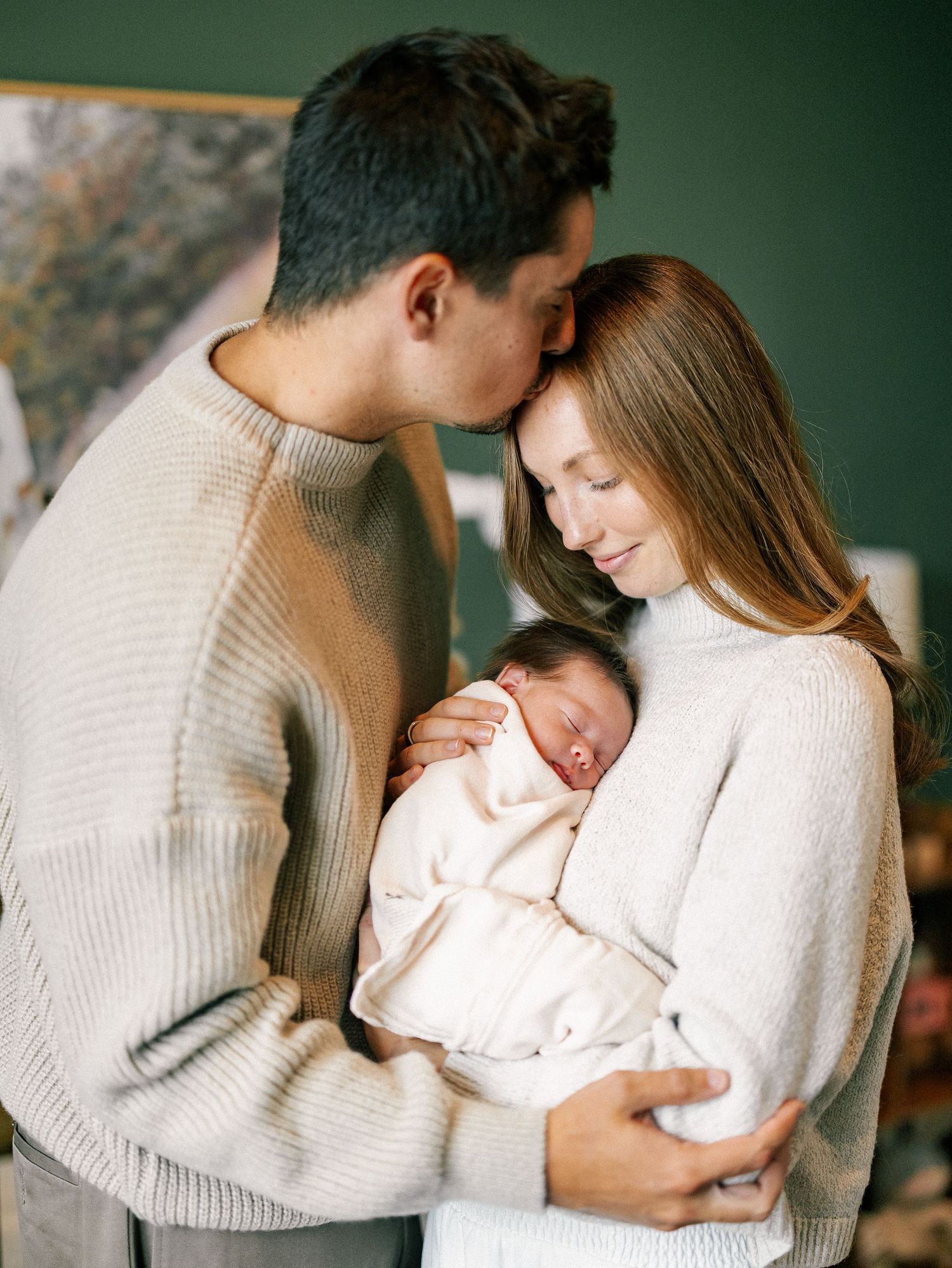 New parents in matching beige sweaters tenderly hold their sleeping newborn baby together indoors.