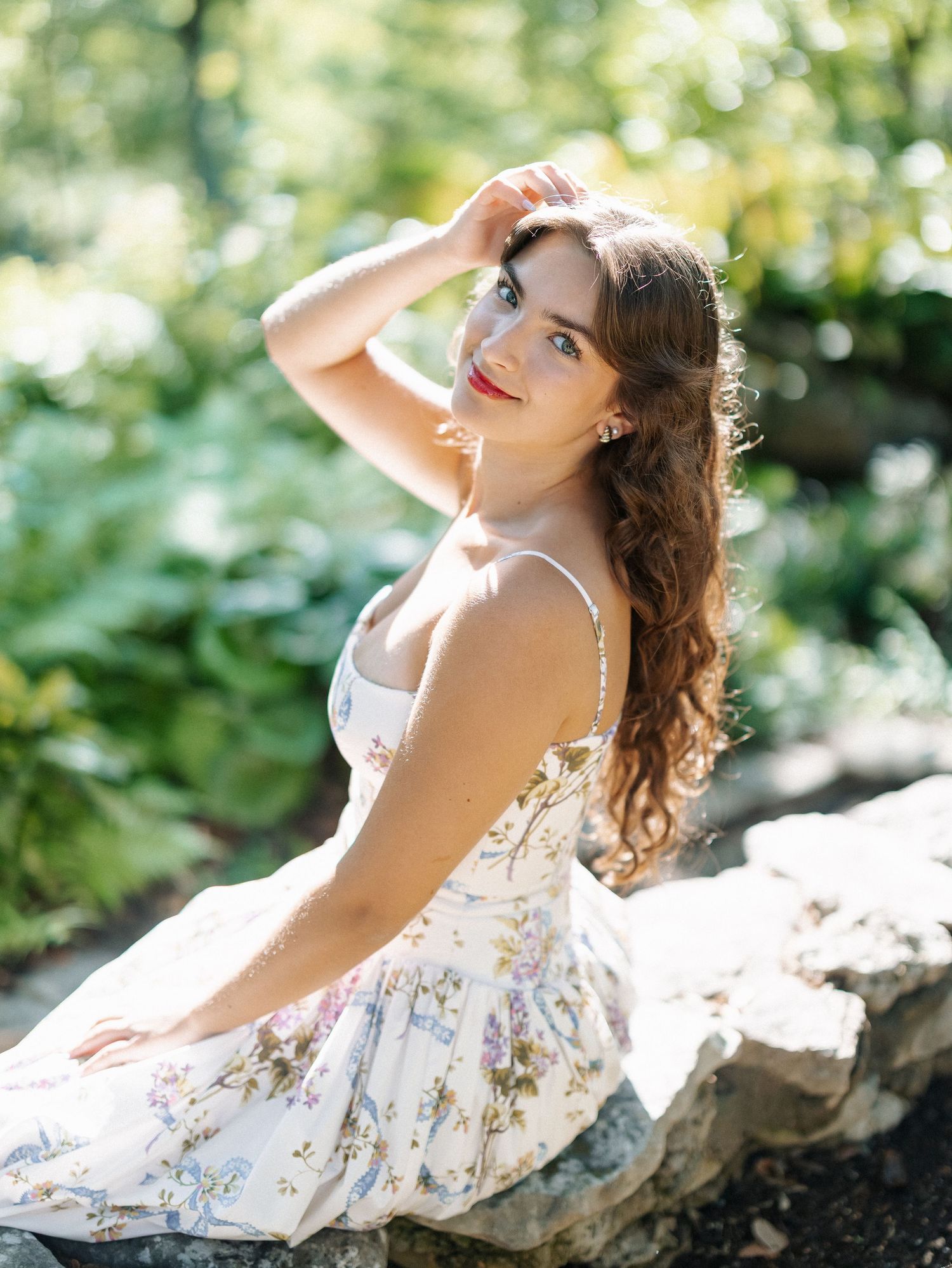 Young woman in floral sundress sitting on rocks in a lush garden, posing with hand in hair.