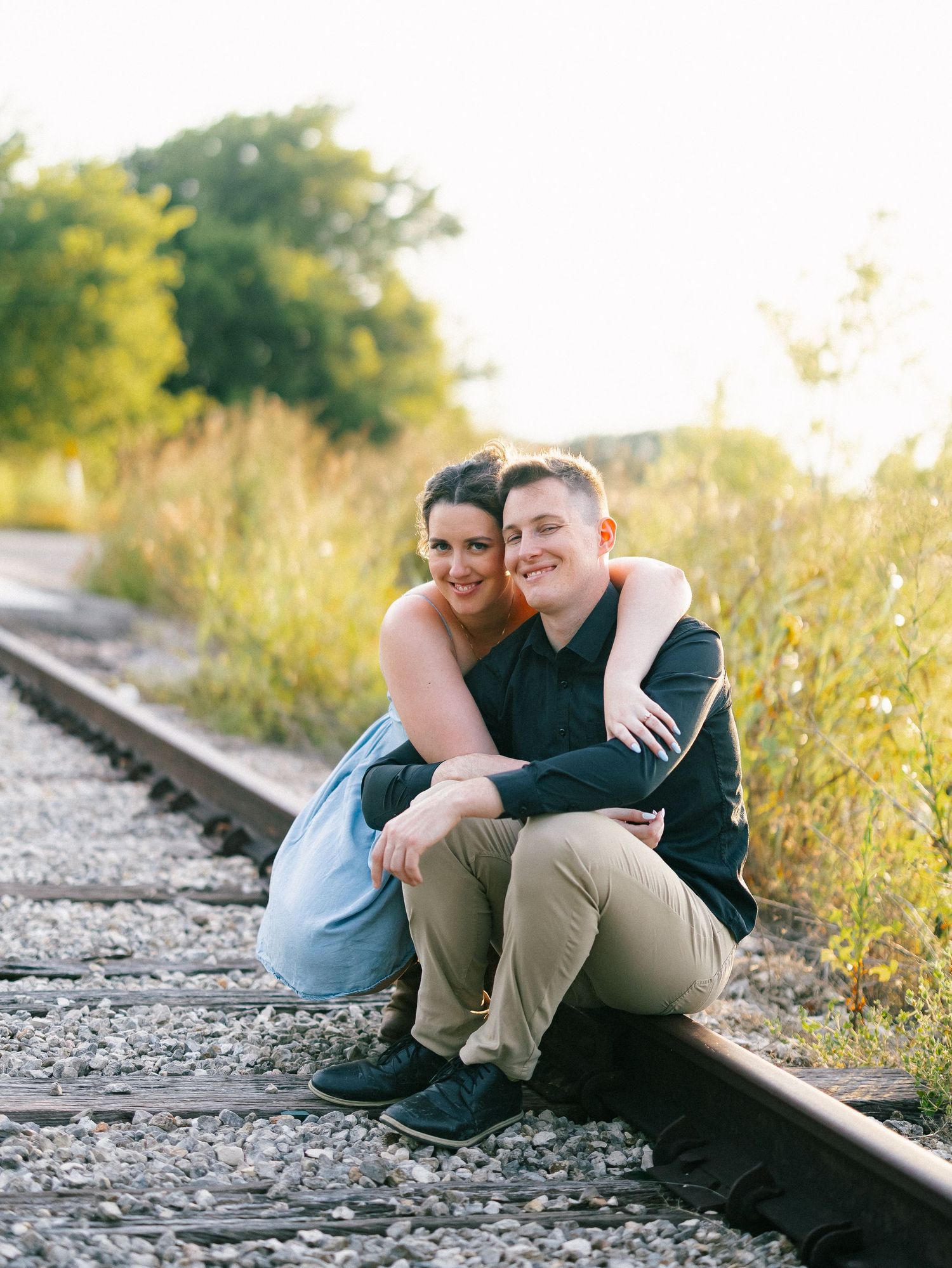 Happy couple embracing on railroad tracks surrounded by golden sunlight during an engagement photo session.