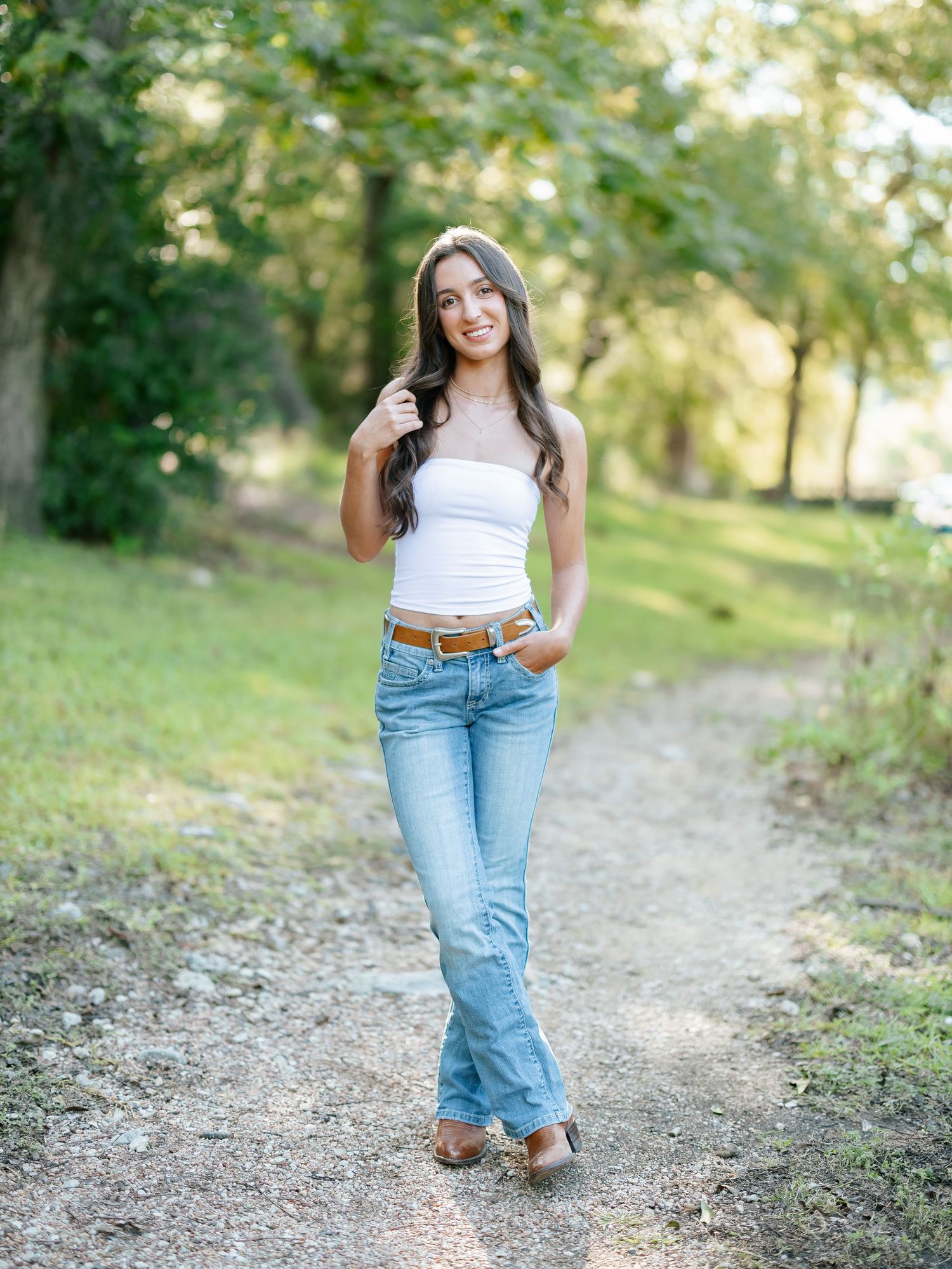 Young woman in white tube top and flare jeans walking on a sunlit gravel path outdoors.