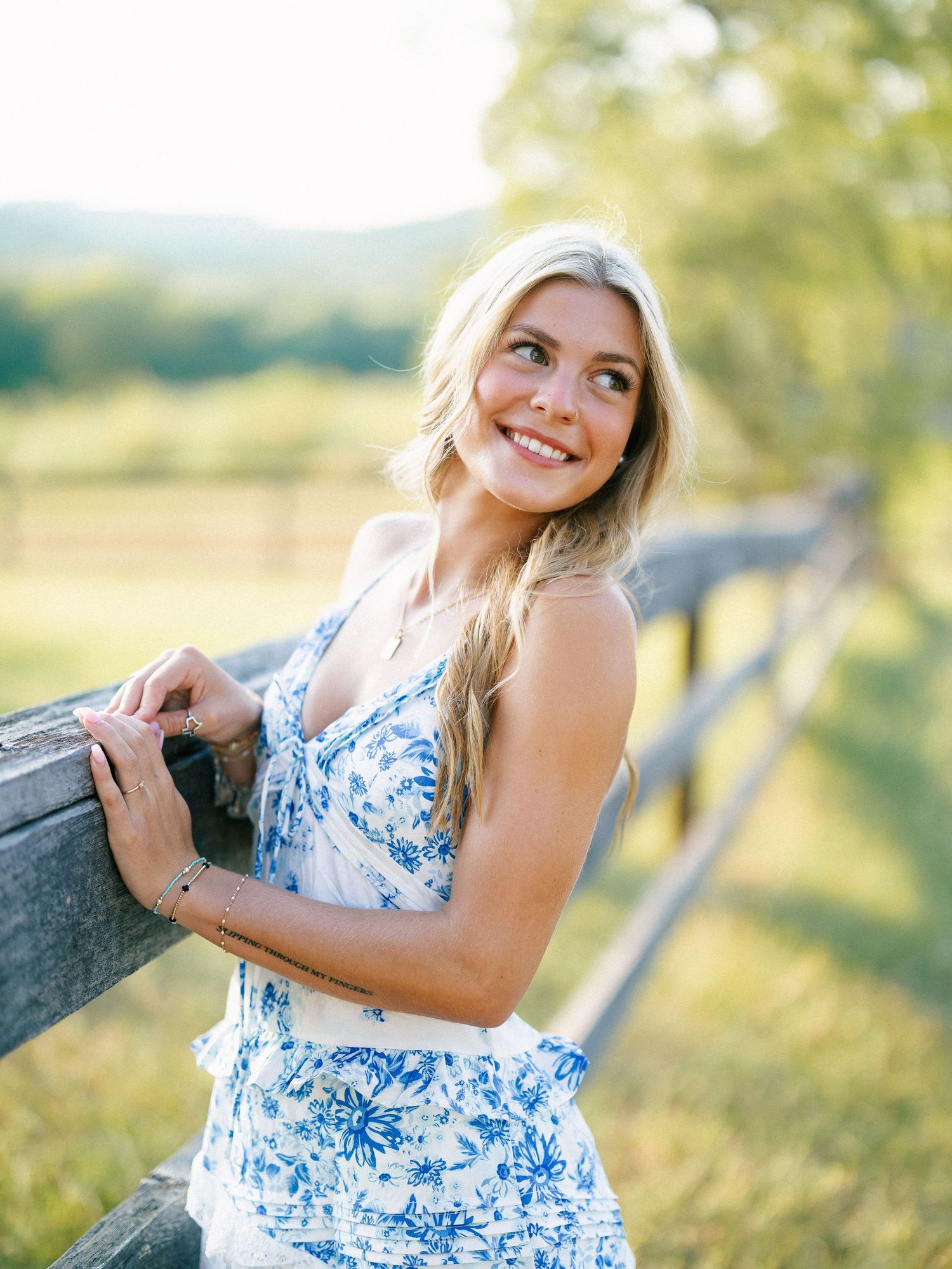 Smiling blonde woman in blue floral dress leaning on rustic wooden fence in sunny countryside setting.
