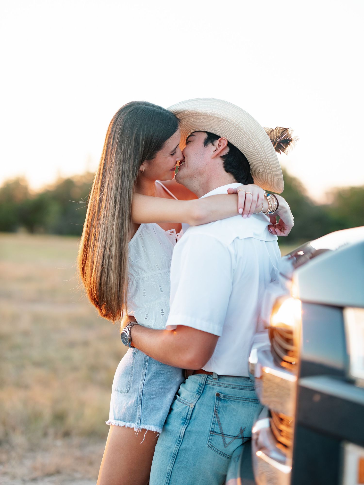 A couple kisses passionately beside a truck at sunset in a rural countryside setting.