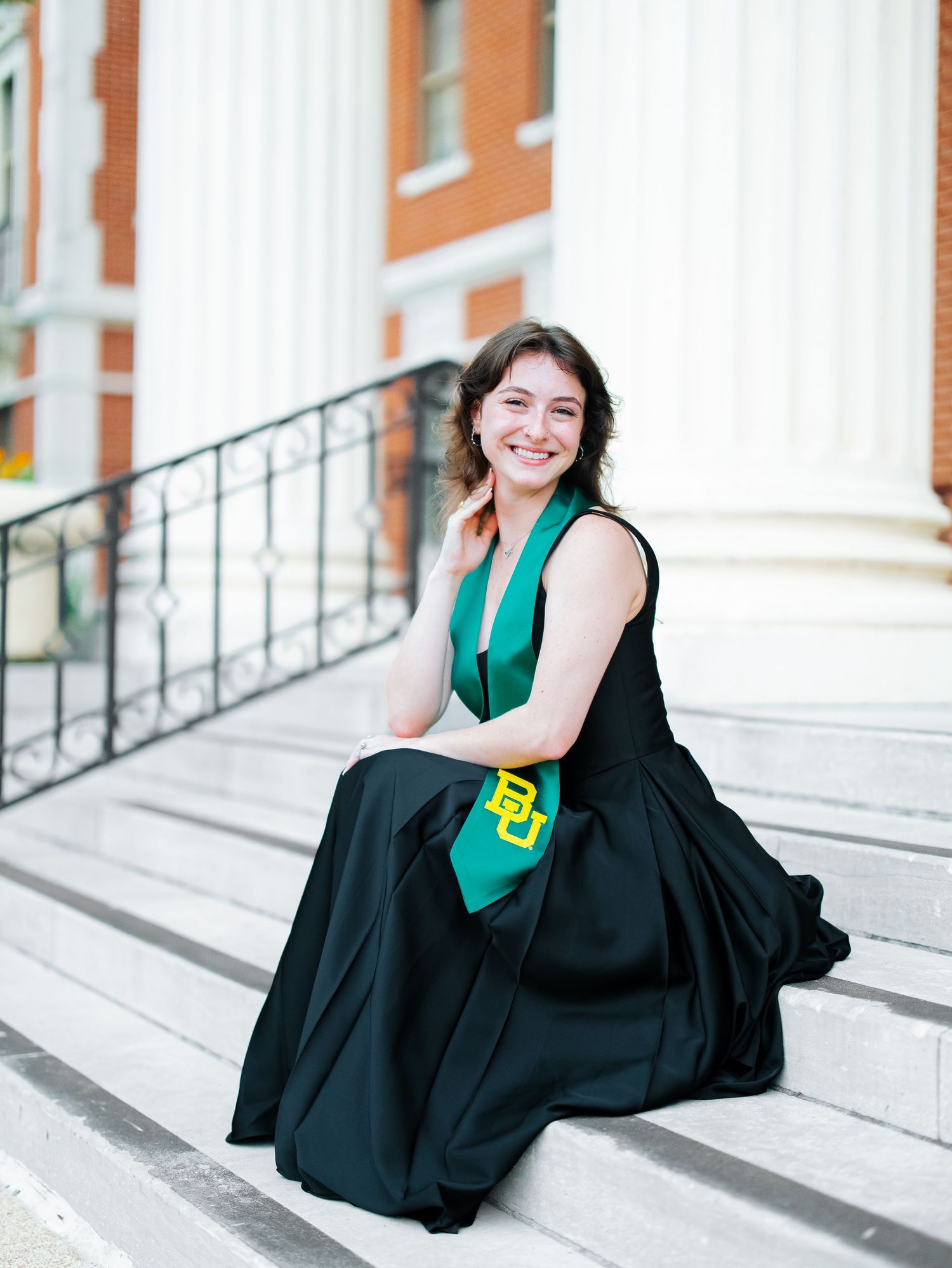 A smiling graduate in a black gown sits on steps wearing a Baylor University green stole.