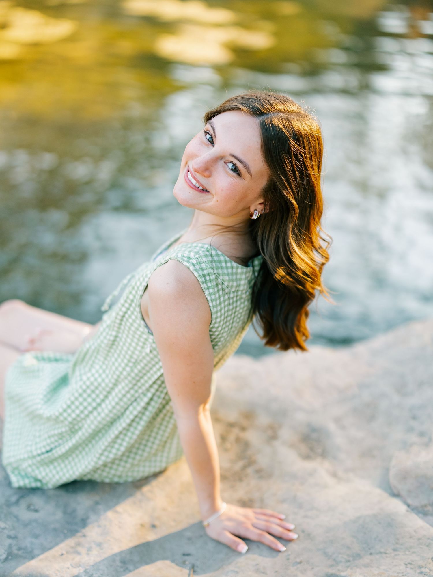 Smiling brunette woman in green gingham dress sitting on rocks near sunlit water.