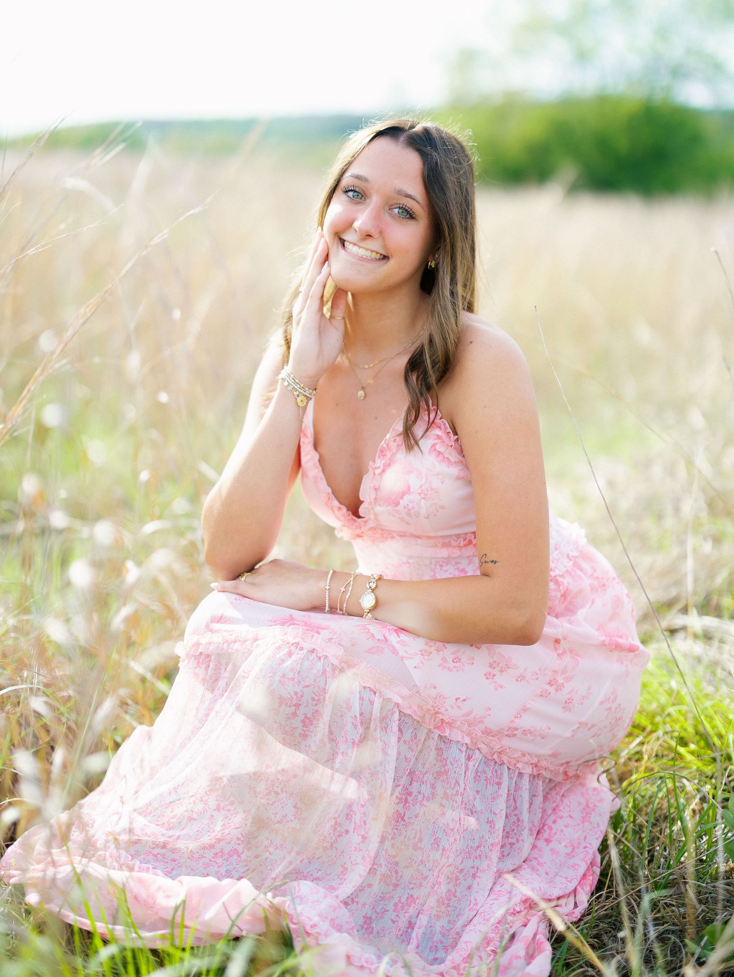 Young woman in pink floral dress sitting in golden field, smiling gracefully during outdoor senior portrait session.