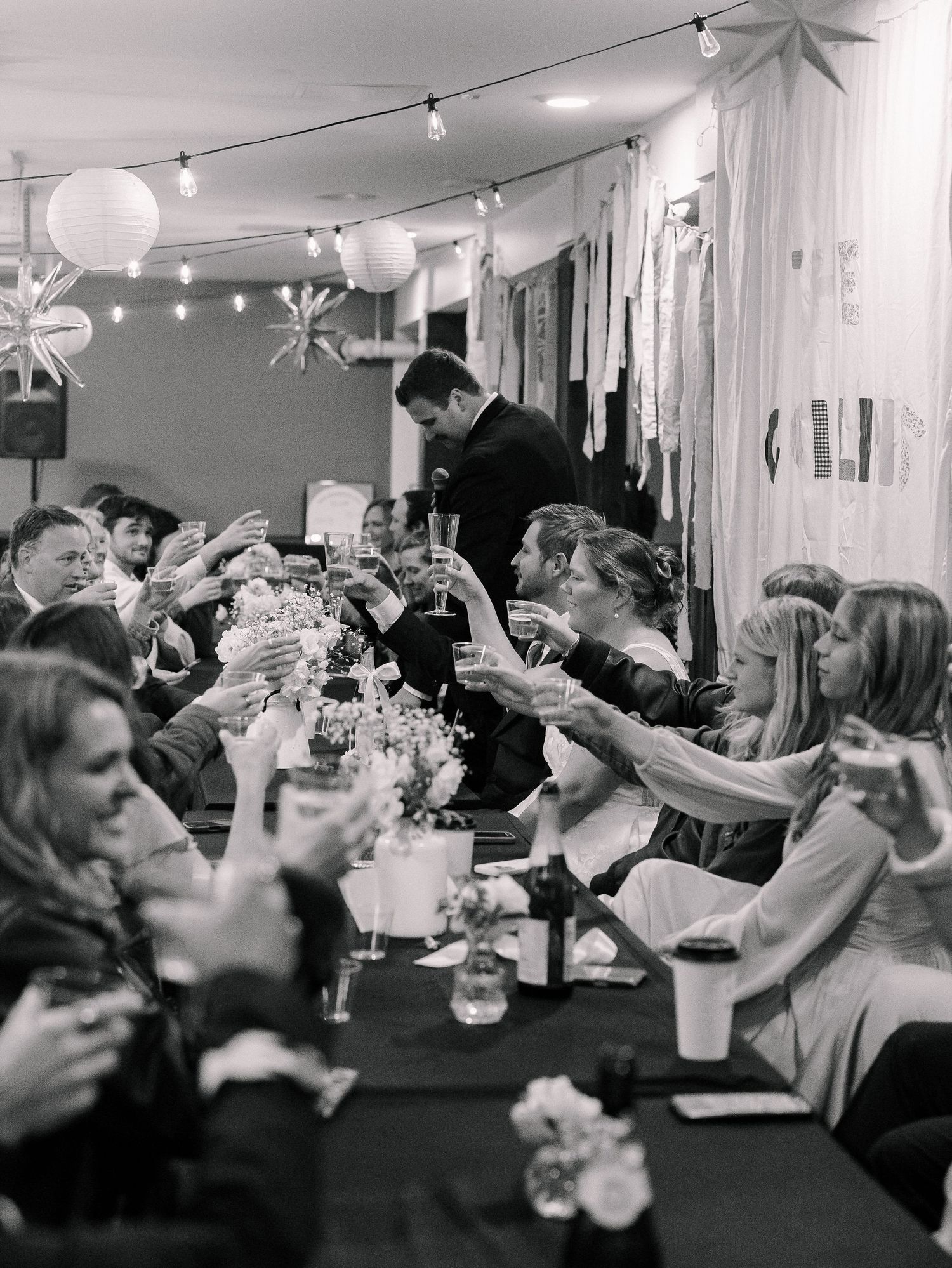 Wedding guests raising glasses in a joyful toast during a lively reception in black and white photography.