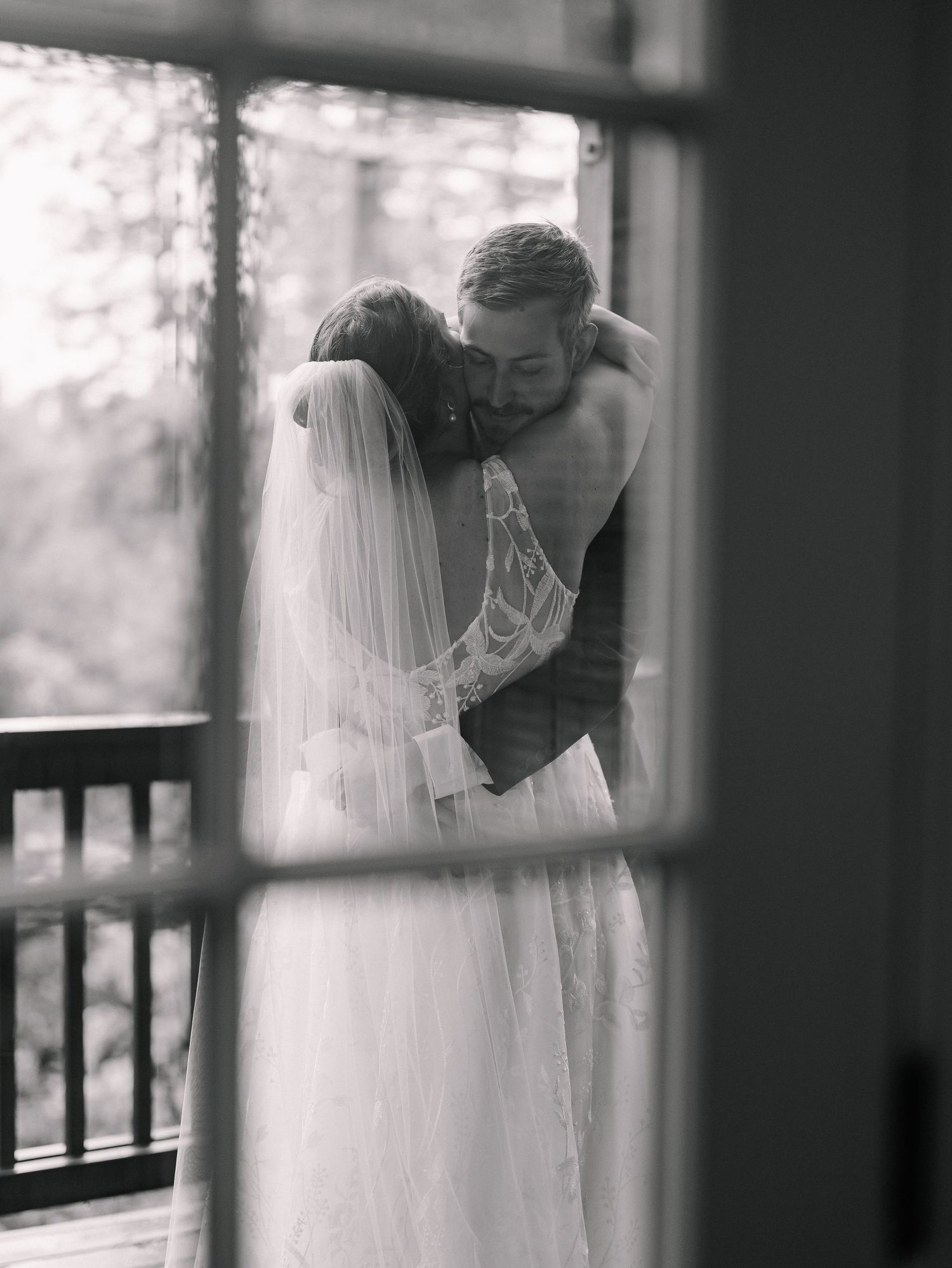 Black and white romantic photo of bride and groom embracing on porch, captured through window with bride in lace gown.