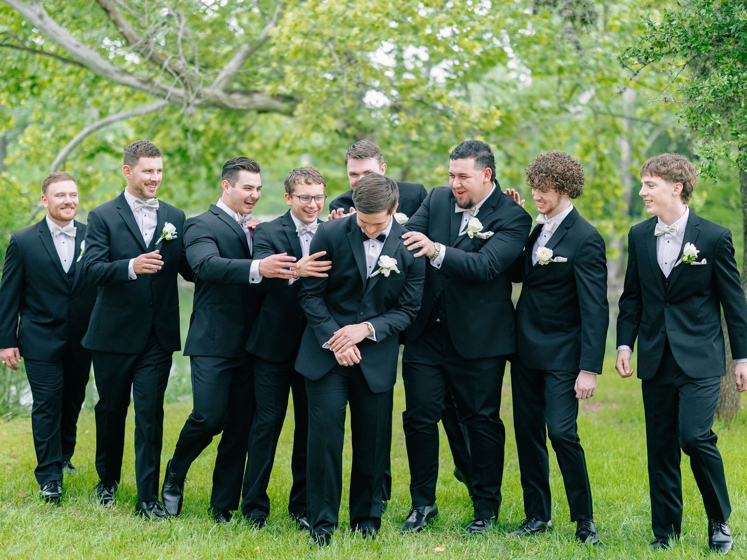 Groomsmen in black suits laugh together outdoors during wedding party photo session.