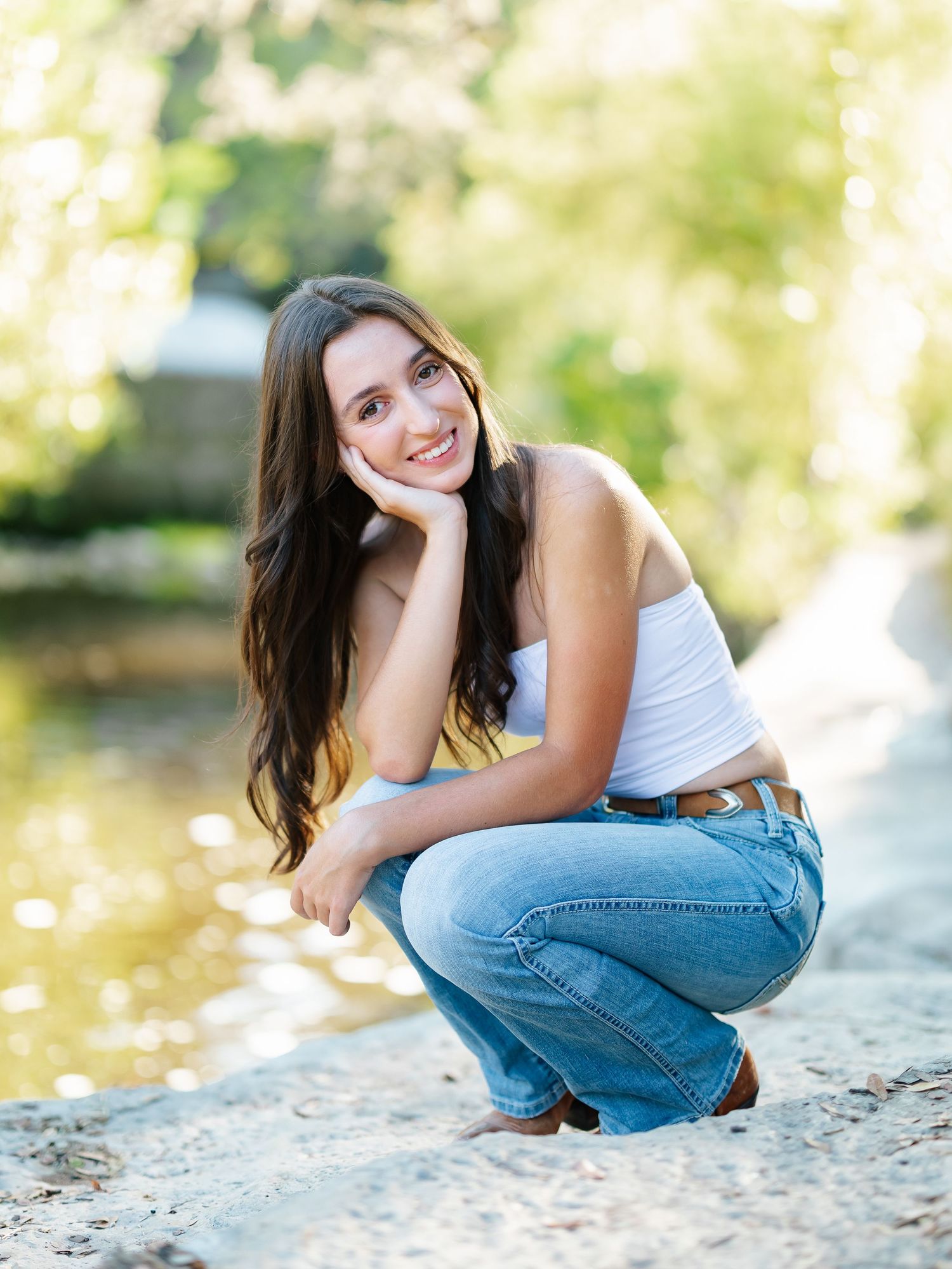 Young woman in white tank top and jeans crouching by a sunlit creek, smiling with hand on chin.