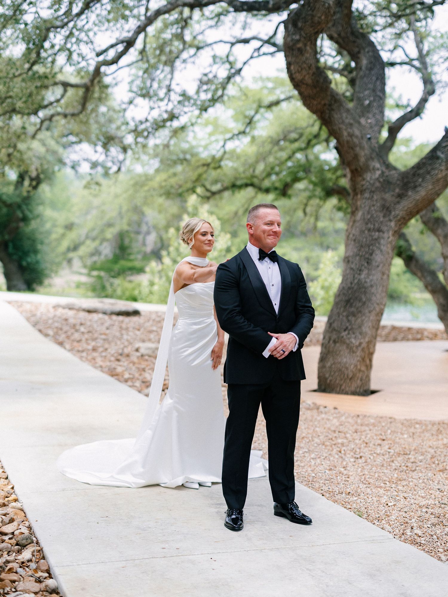 Bride in white halter gown stands with groom in black tuxedo during outdoor first look under oak trees.