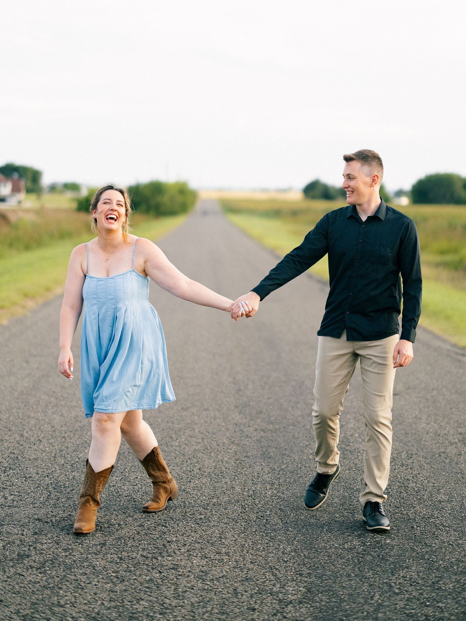 A laughing couple holds hands while walking down a rural road, woman in blue dress and cowboy boots, man in black shirt.