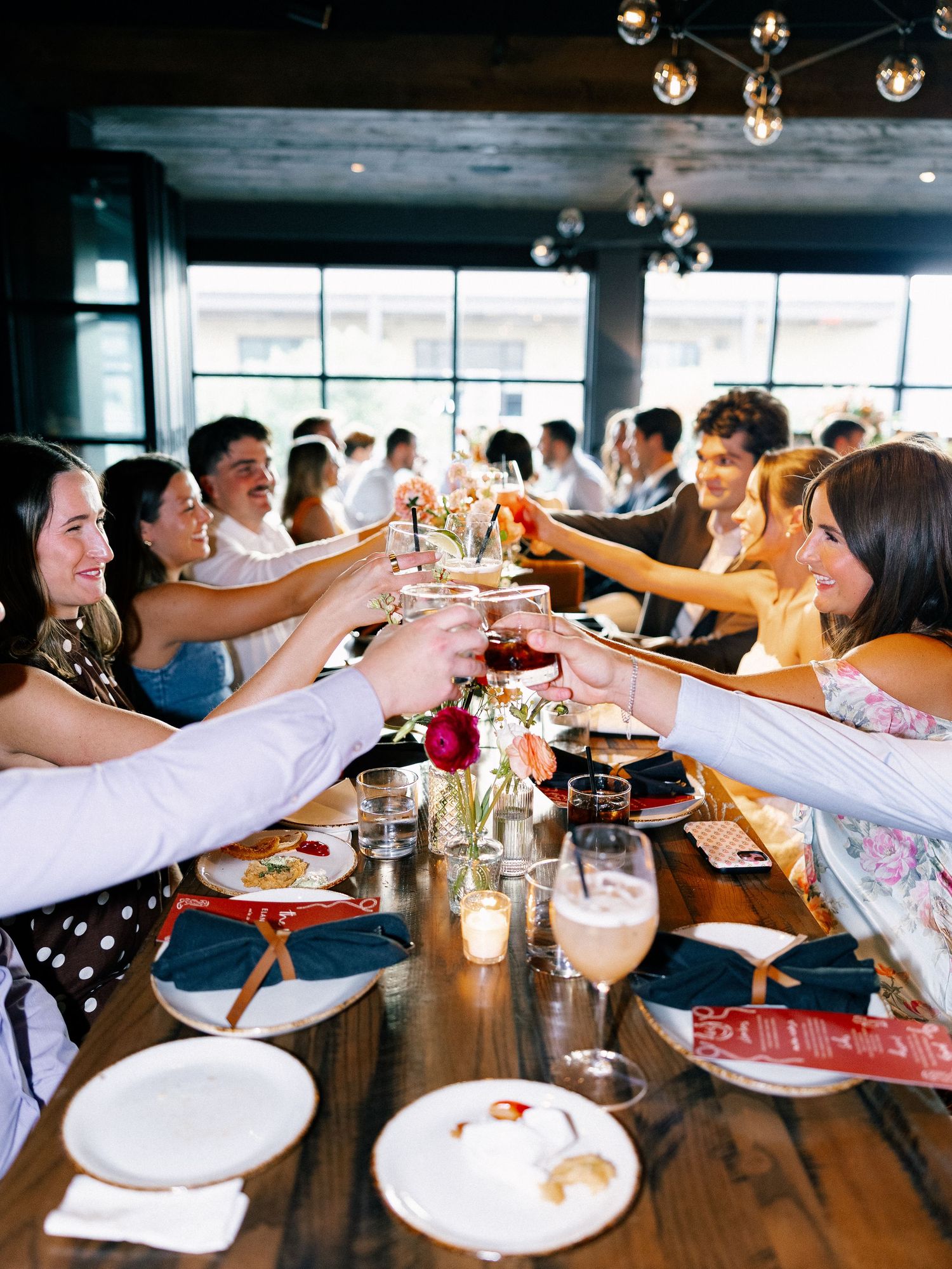 Guests raising glasses in a toast at a long wooden table during a celebratory dinner party in a modern restaurant.