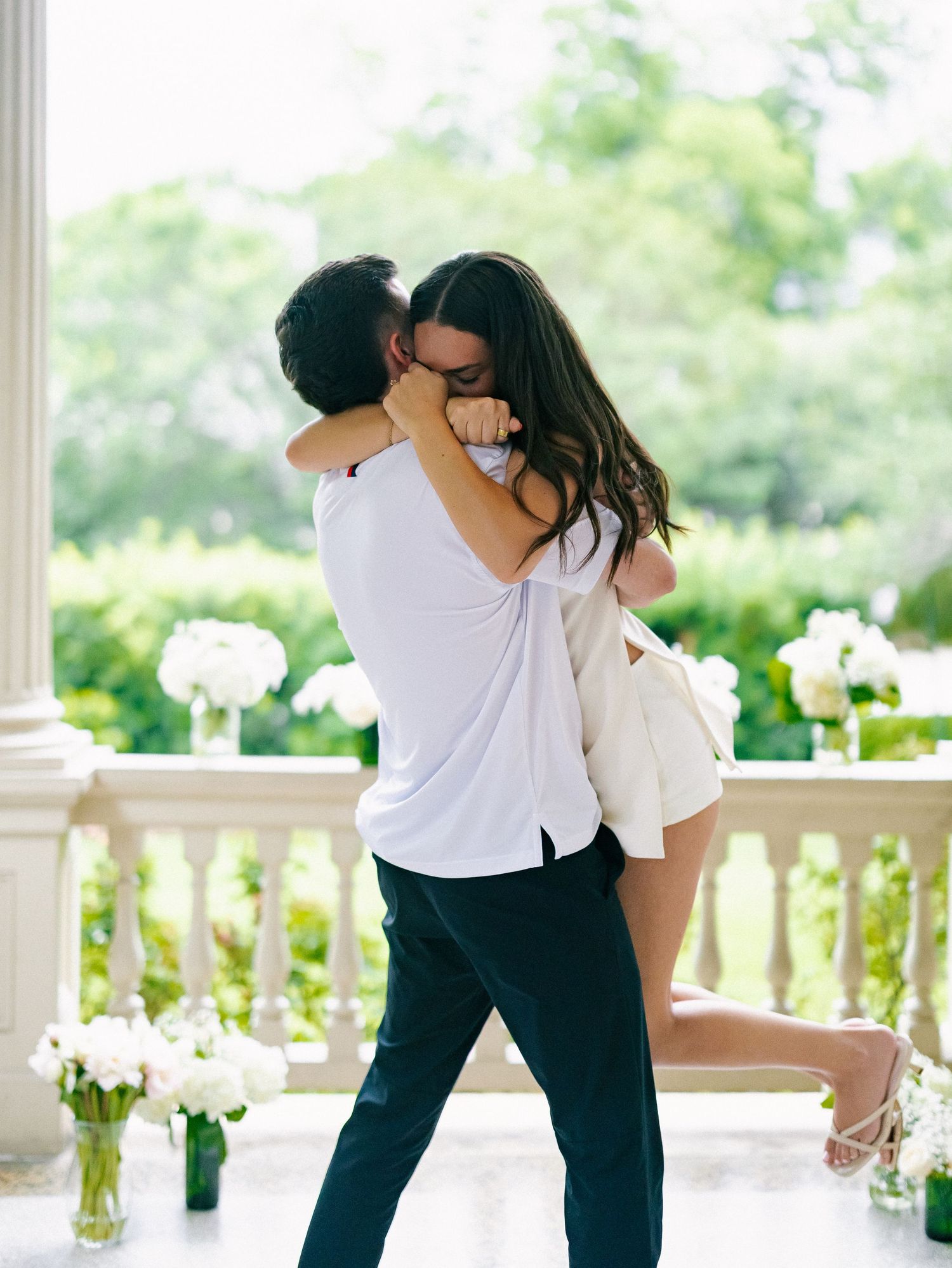 Couple sharing romantic embrace on elegant white columned porch surrounded by white floral arrangements.