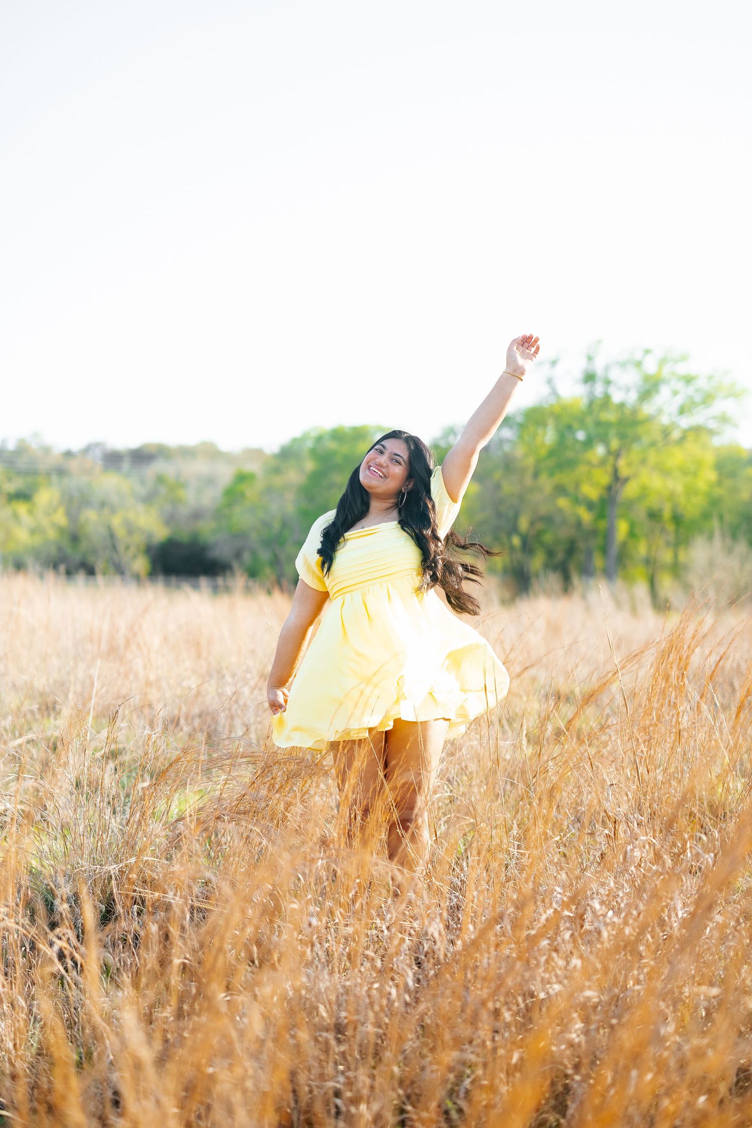 Young woman in yellow dress dancing joyfully with arm raised in golden field at sunset.