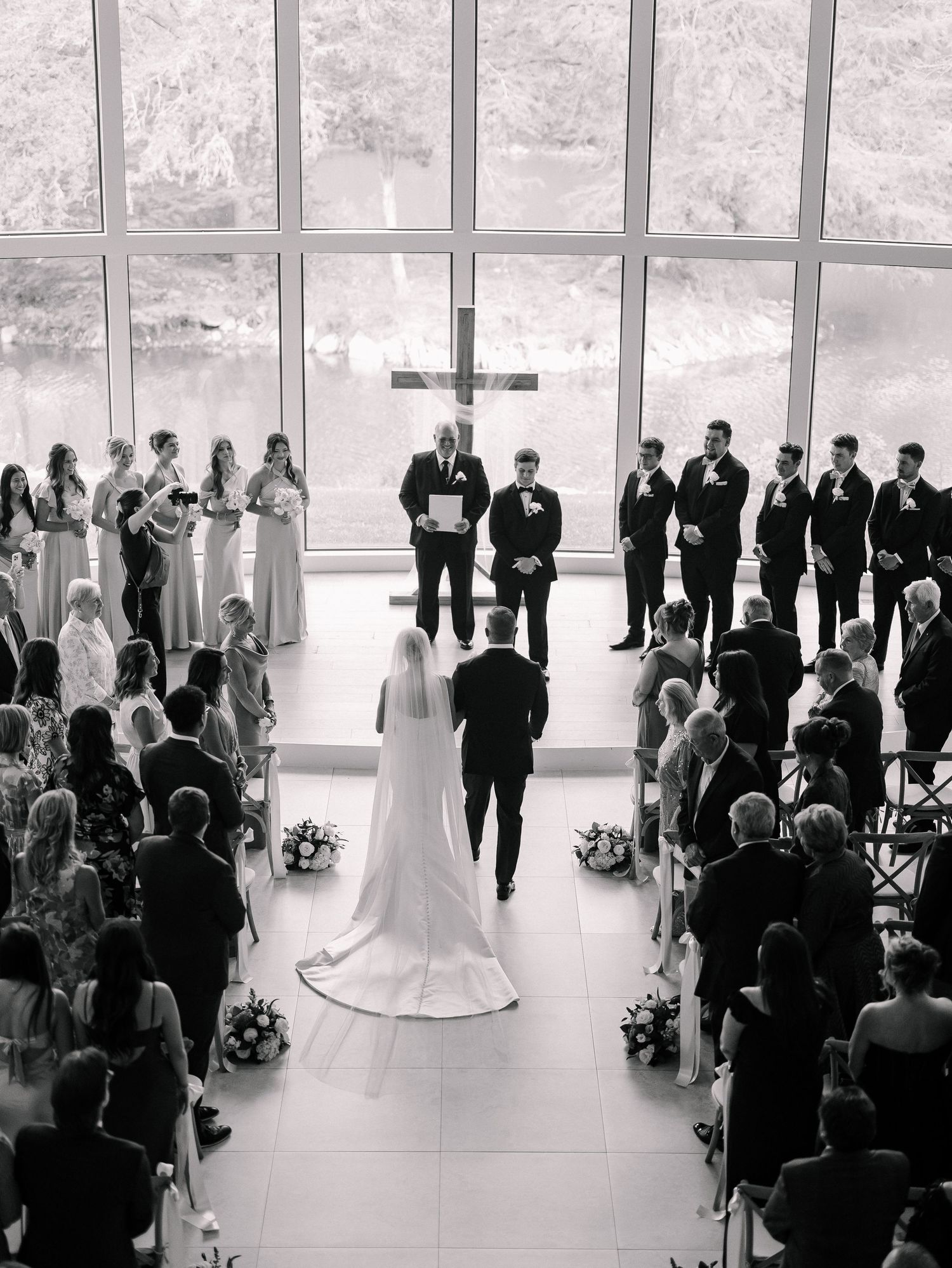 Aerial black and white photo of bride walking down aisle toward groom at chapel altar with floor-to-ceiling windows.