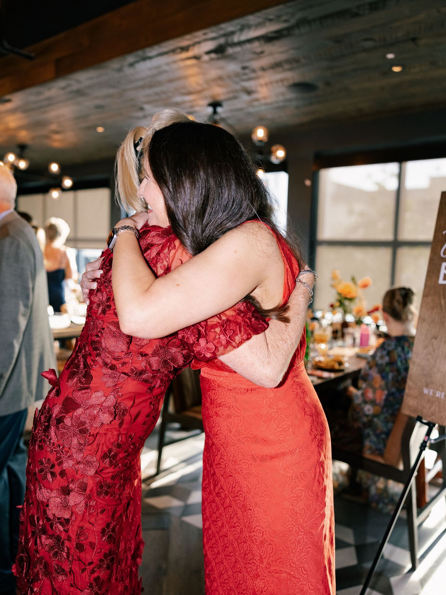 Two women in red lace dresses share an emotional hug at a wedding reception.