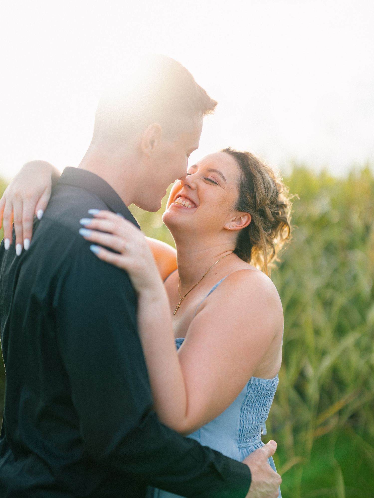 A joyful couple embraces outdoors at golden hour, the woman laughing as they share a romantic moment in a sunlit field.