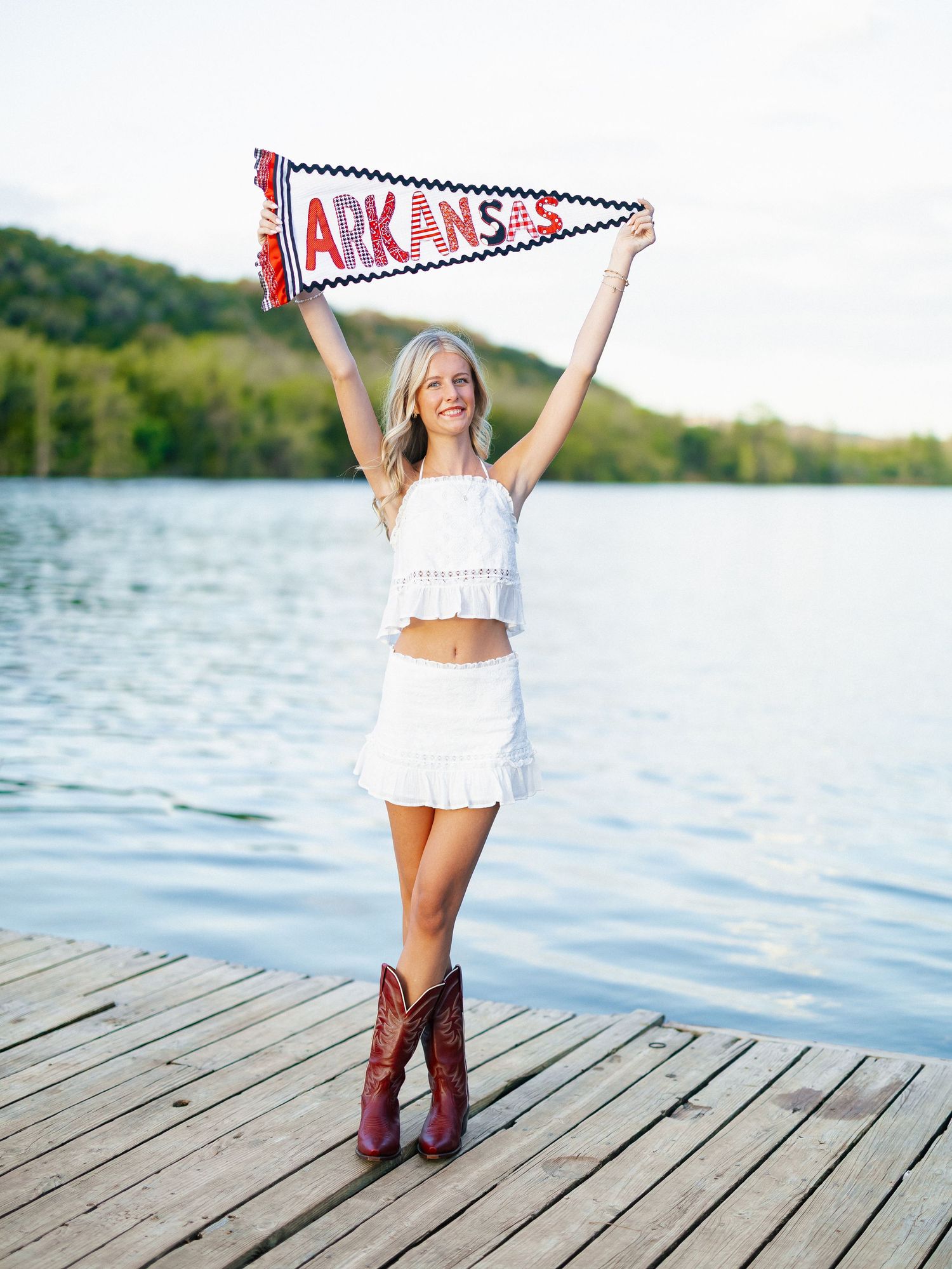 Blonde woman in white crop top and skirt holding Arkansas pennant flag on lake dock wearing cowboy boots.