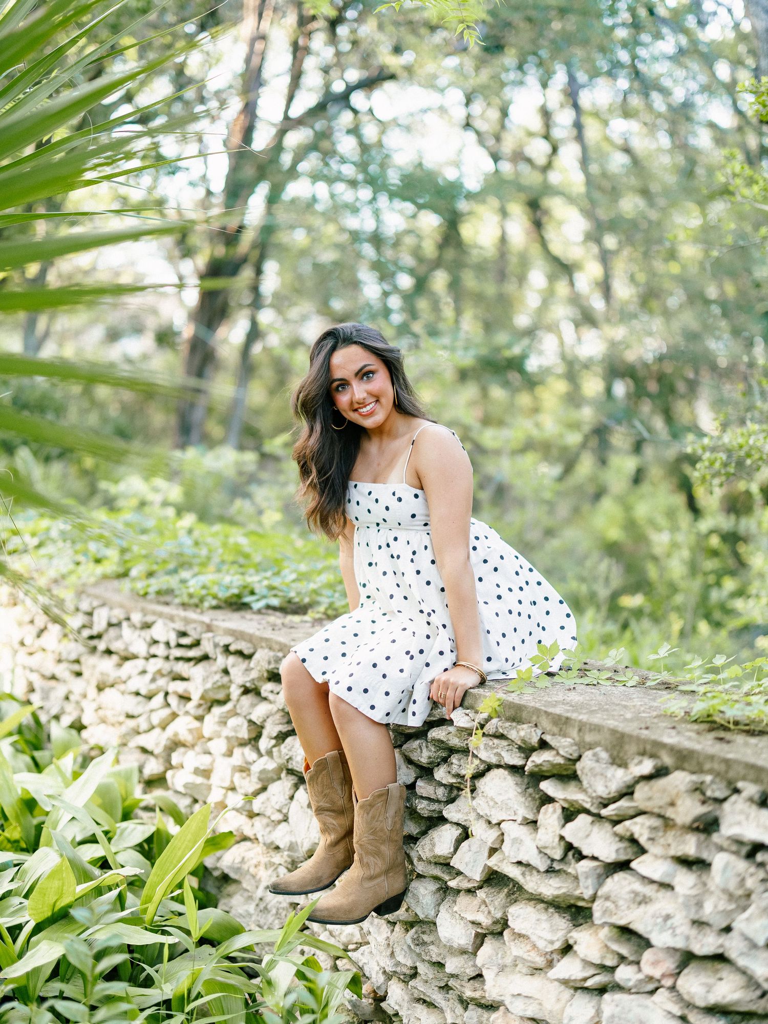 Young woman in white polka dot dress and brown boots sitting on a stone wall surrounded by lush green trees.