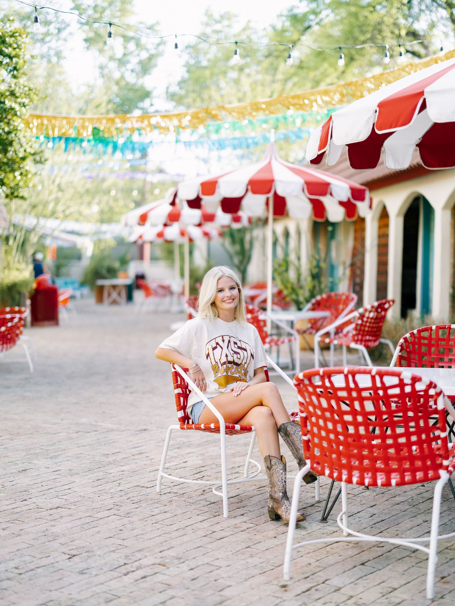 Smiling blonde woman in a graphic tee, denim shorts, and cowboy boots sitting in a red chair at a colorful outdoor patio.