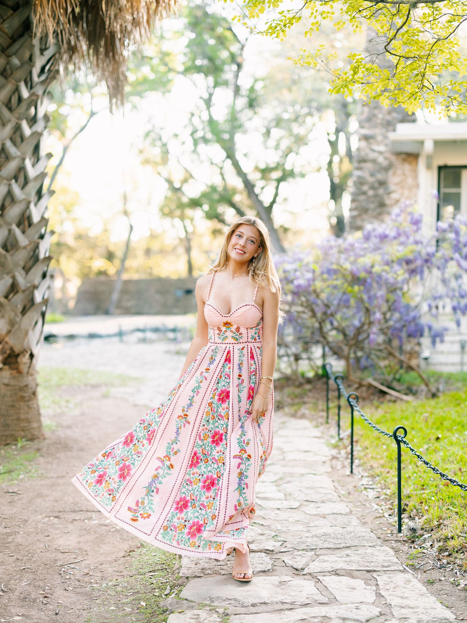 Woman in floral pink maxi dress walking on stone path surrounded by blooming trees and greenery.