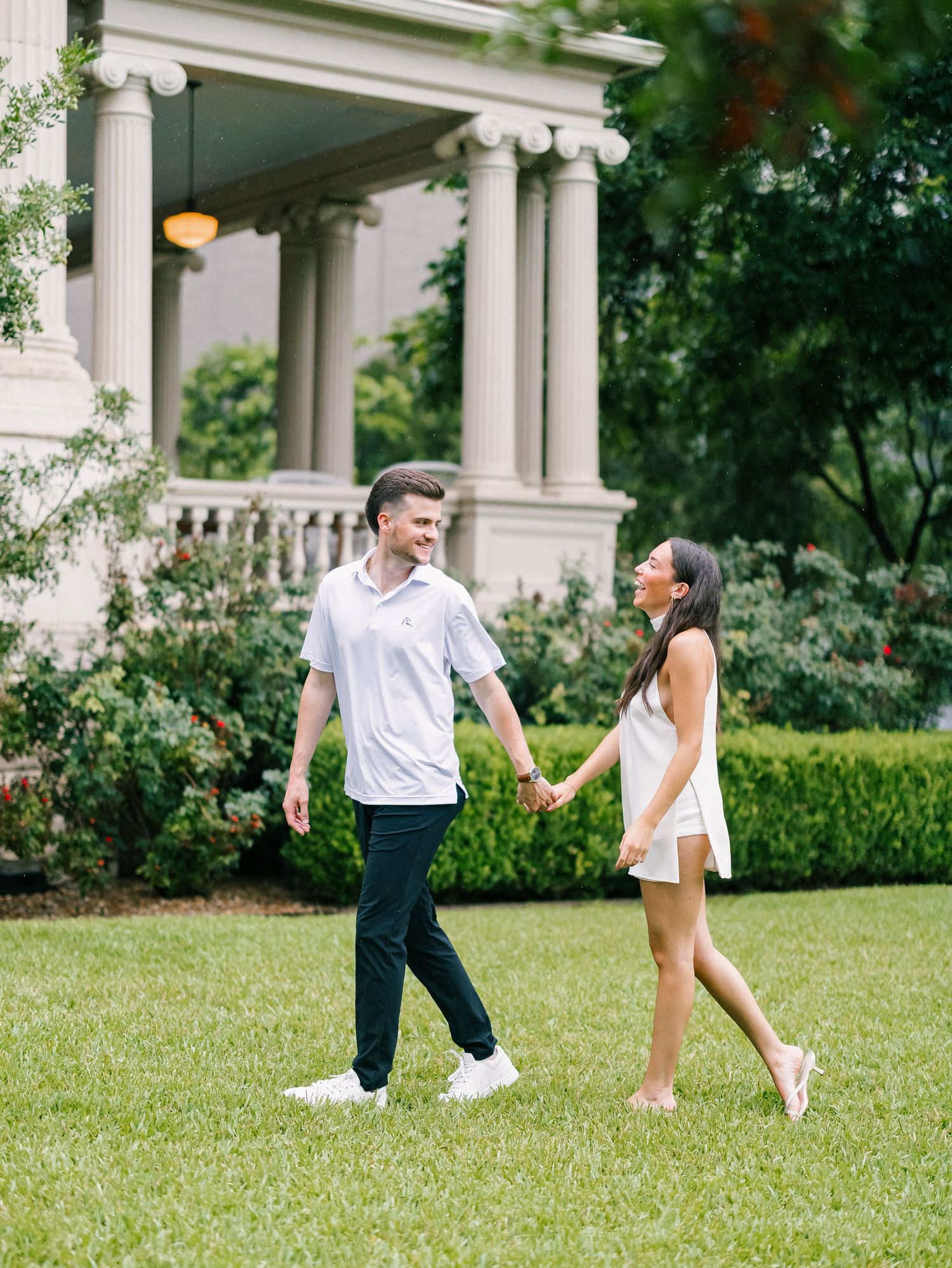 A couple holds hands and walks on green grass in front of an elegant white columned mansion surrounded by lush hedges.