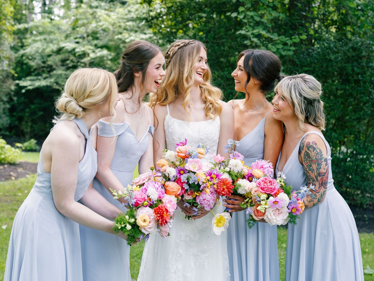 Bride and three bridesmaids in blue dresses laugh together holding colorful bridal bouquets outdoors.