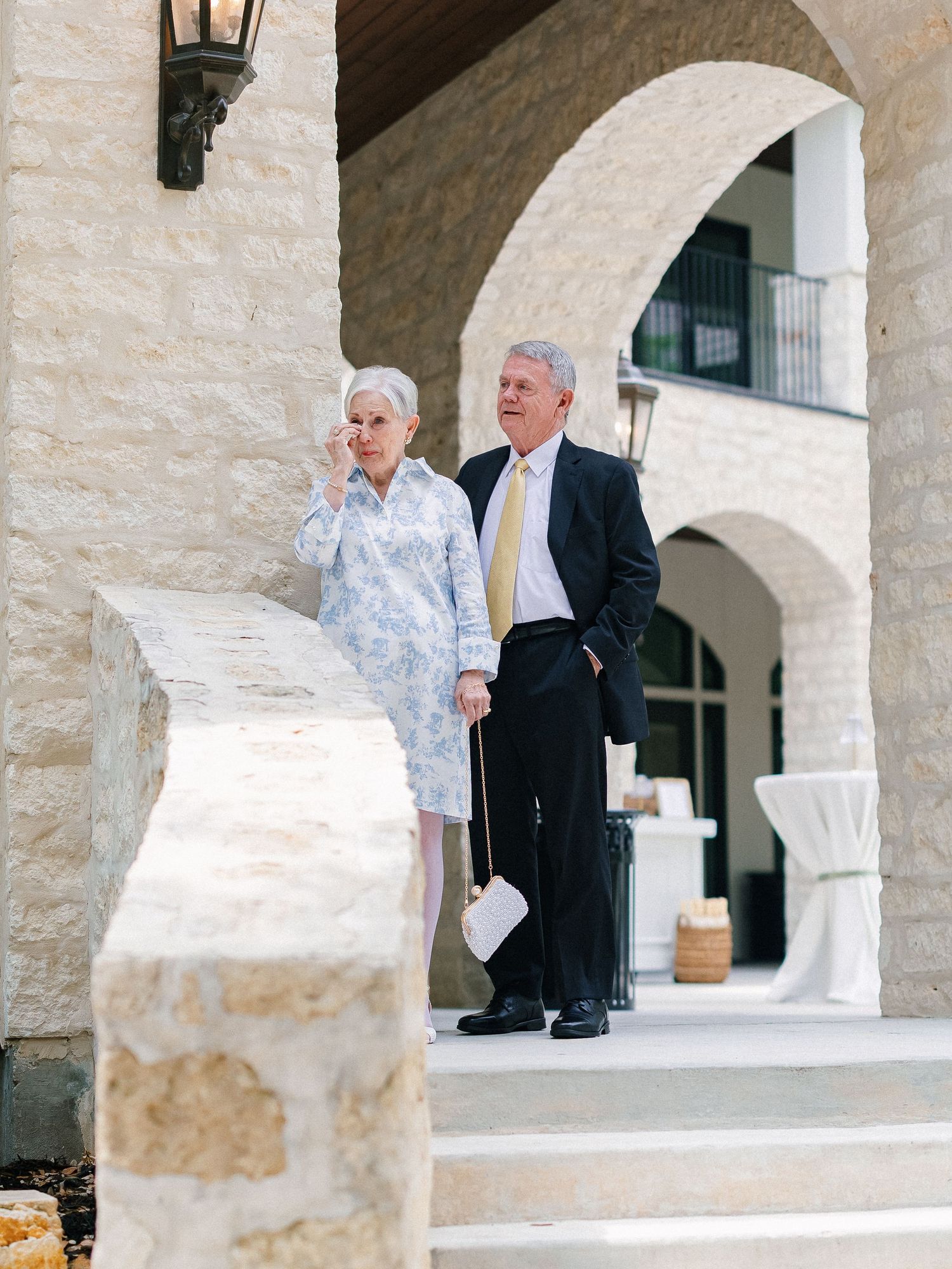 Elderly couple standing outside a stone building with arched doorways, woman in floral dress, man in dark suit with yellow tie.