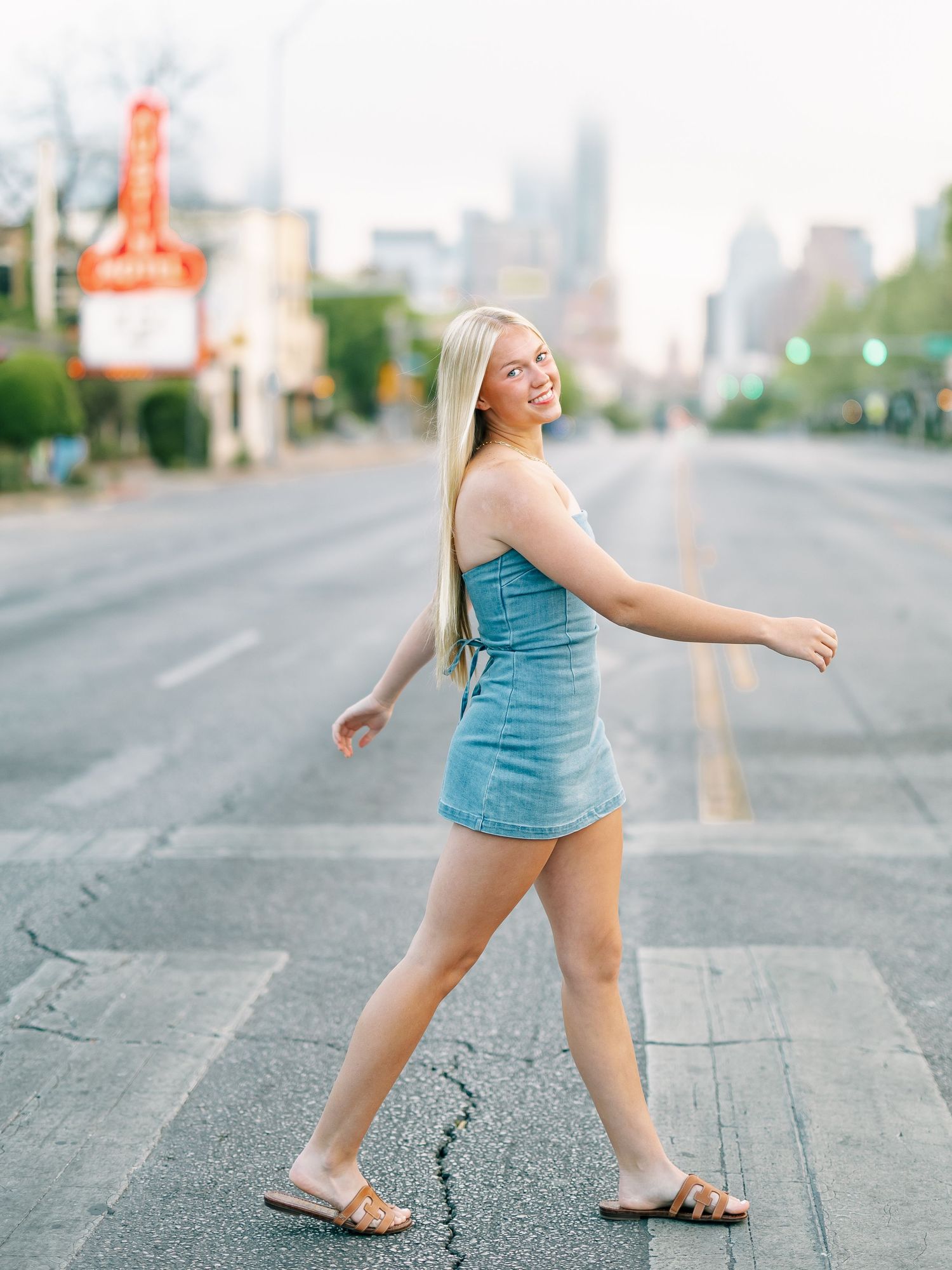 Blonde woman in denim dress crossing empty city street near Austin Motel sign at dusk.