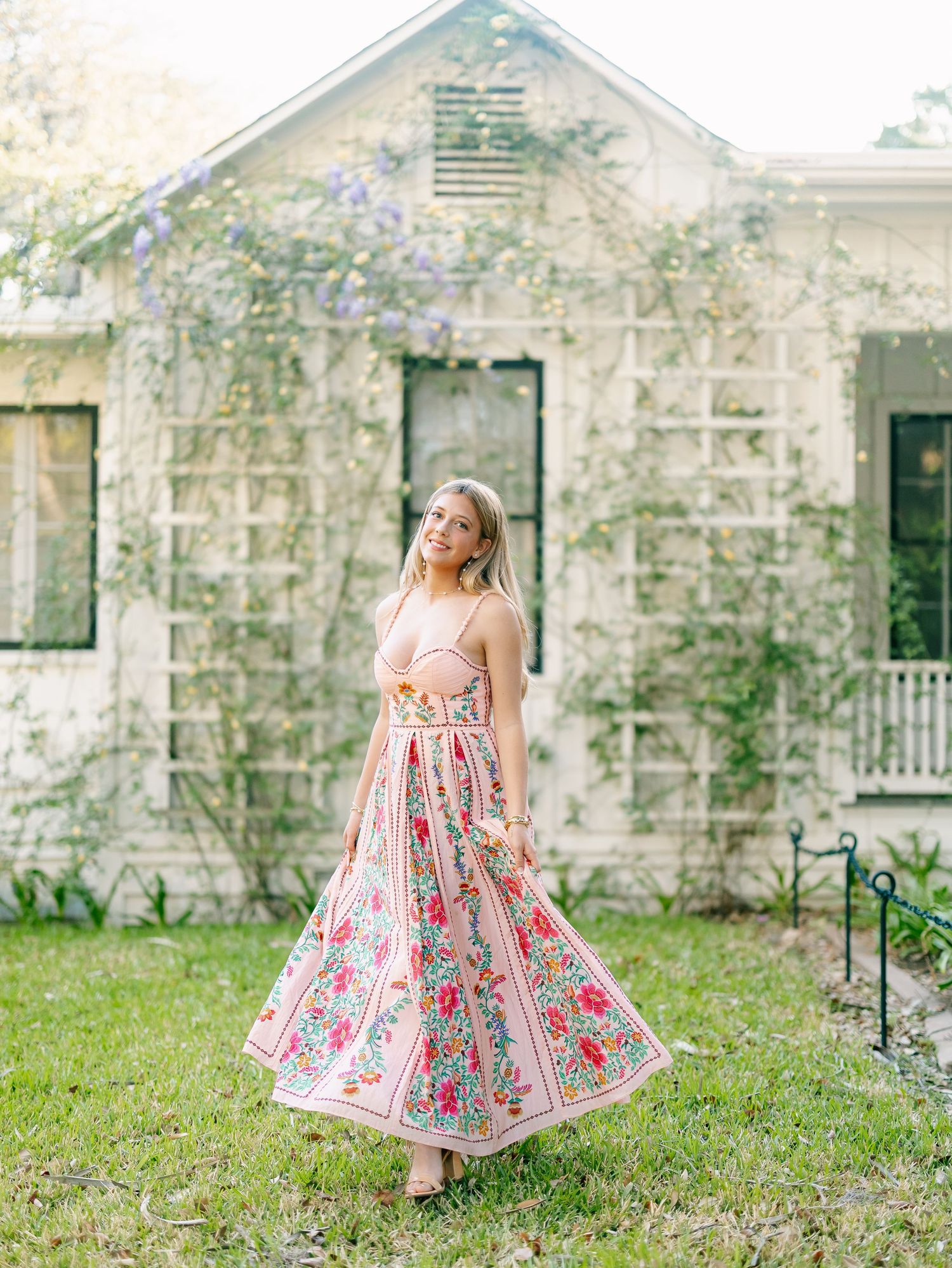 Blonde woman in pink floral maxi dress standing on green lawn before vintage cottage.