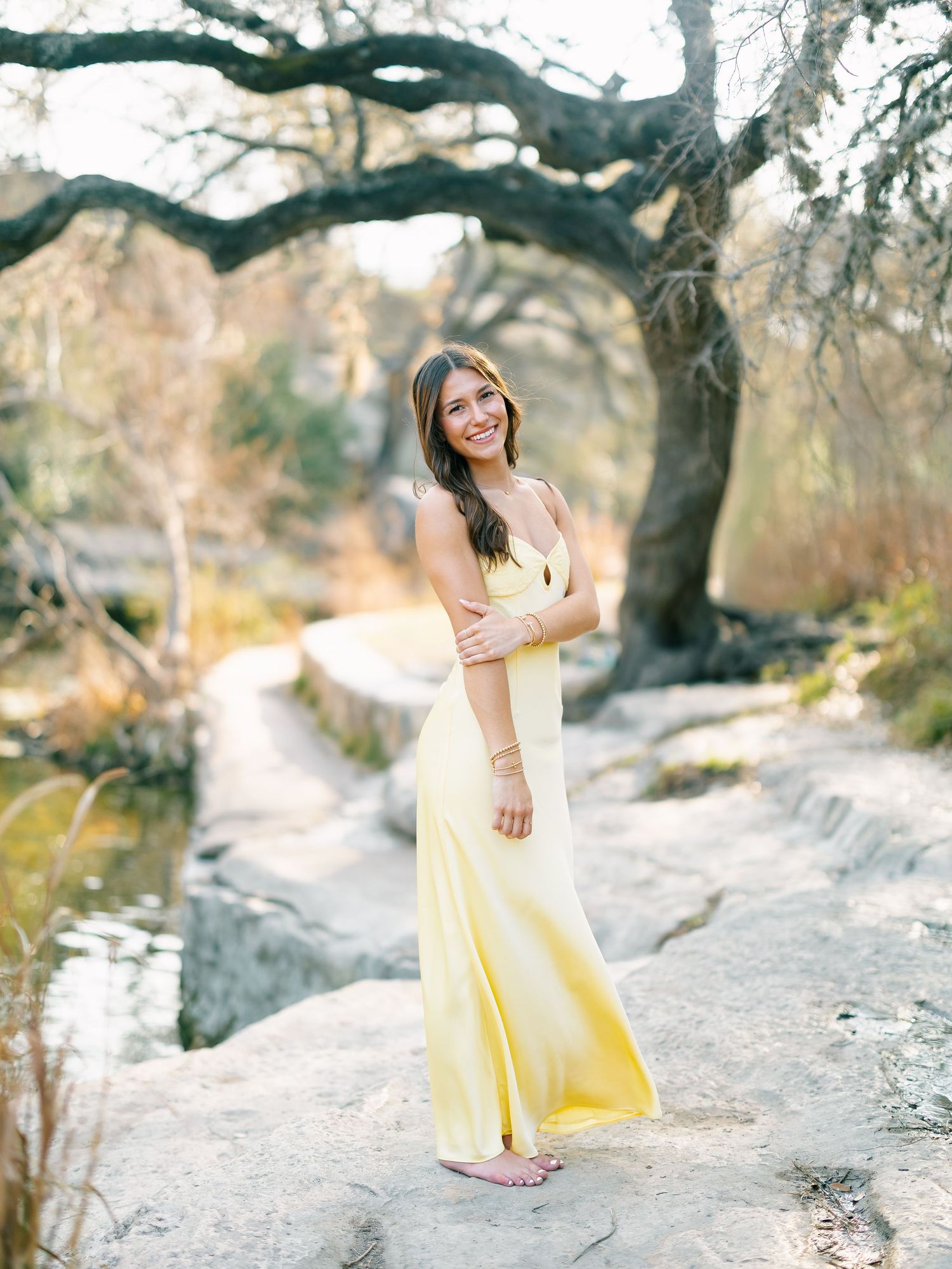 Young woman in yellow maxi dress stands on rocky creek bed beneath twisted oak tree.