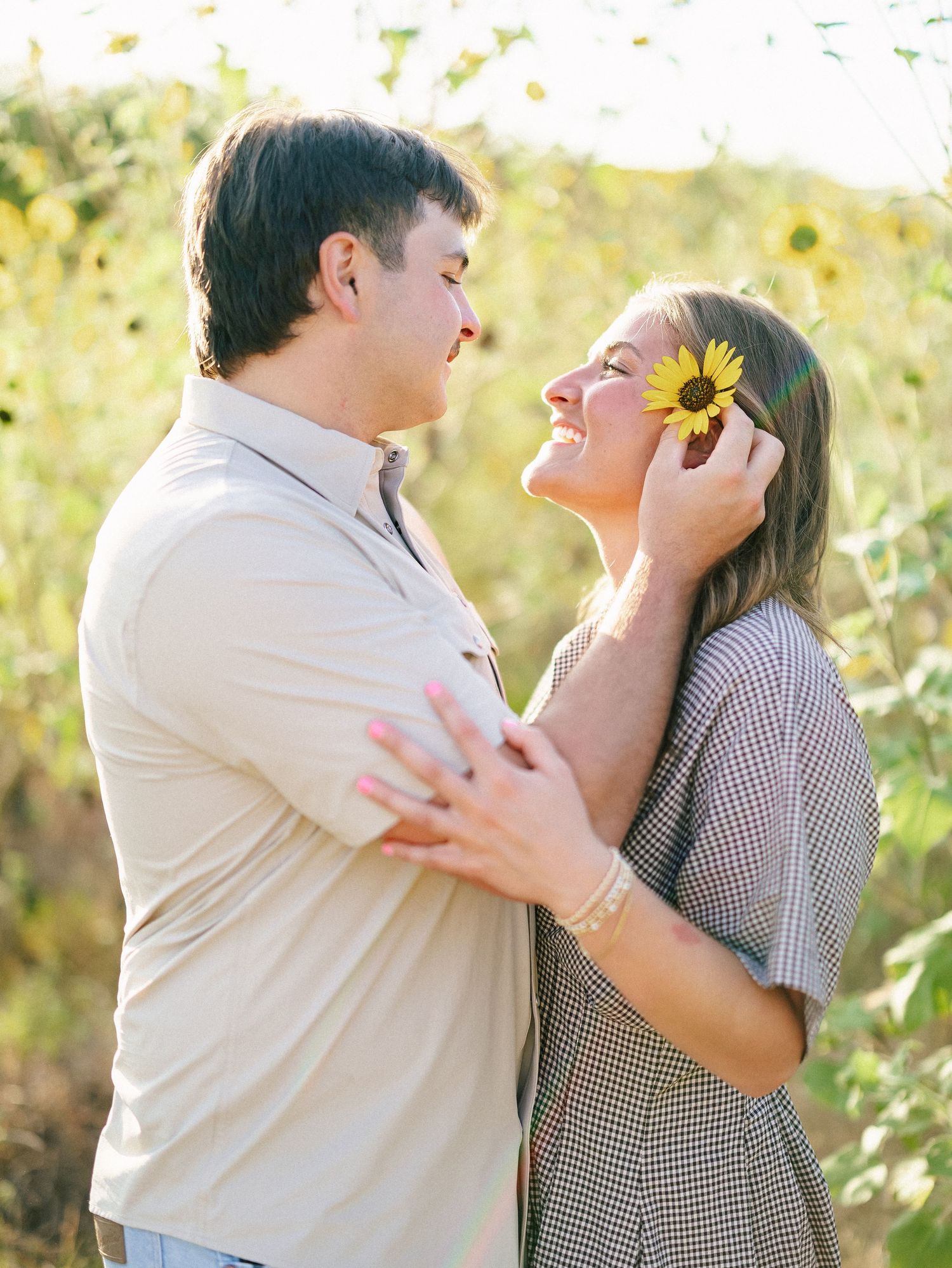 A couple shares a romantic moment in a sunflower field, as the woman holds a sunflower up to her face while smiling.