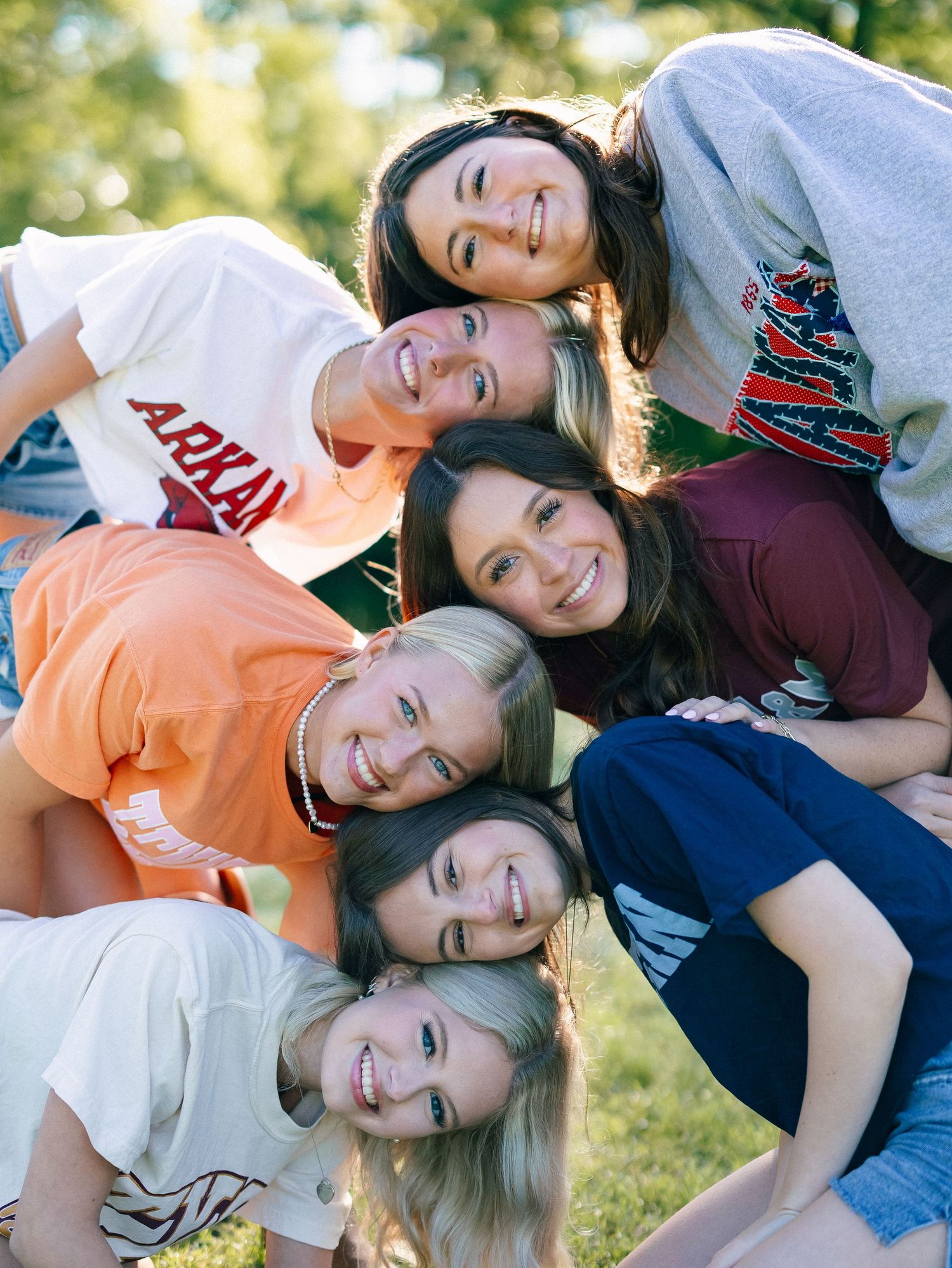Six smiling young women stacking their heads together in a fun pyramid pose outdoors on green grass.
