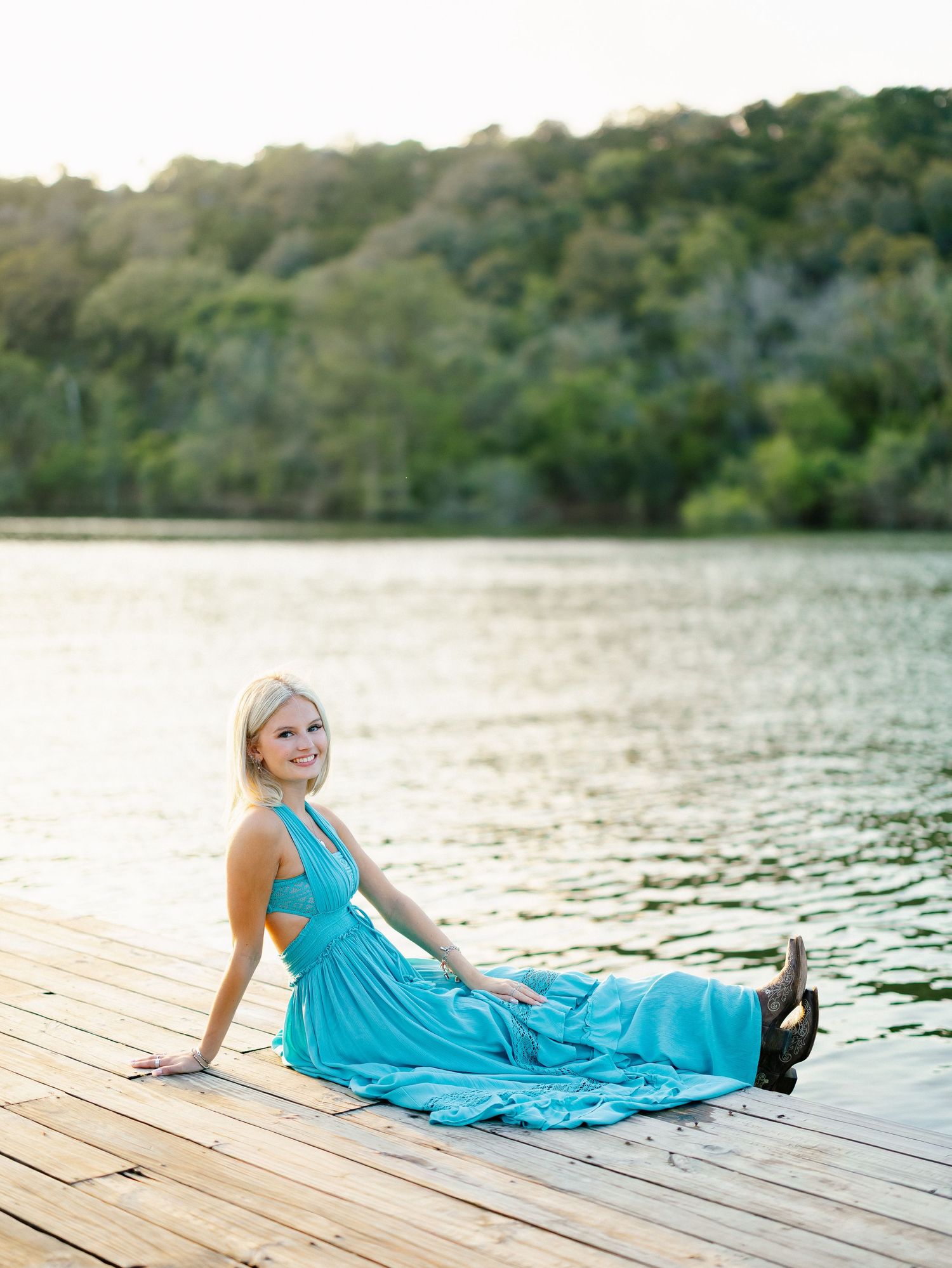 Blonde senior girl in a teal maxi dress sits gracefully on a wooden dock overlooking a serene lake.