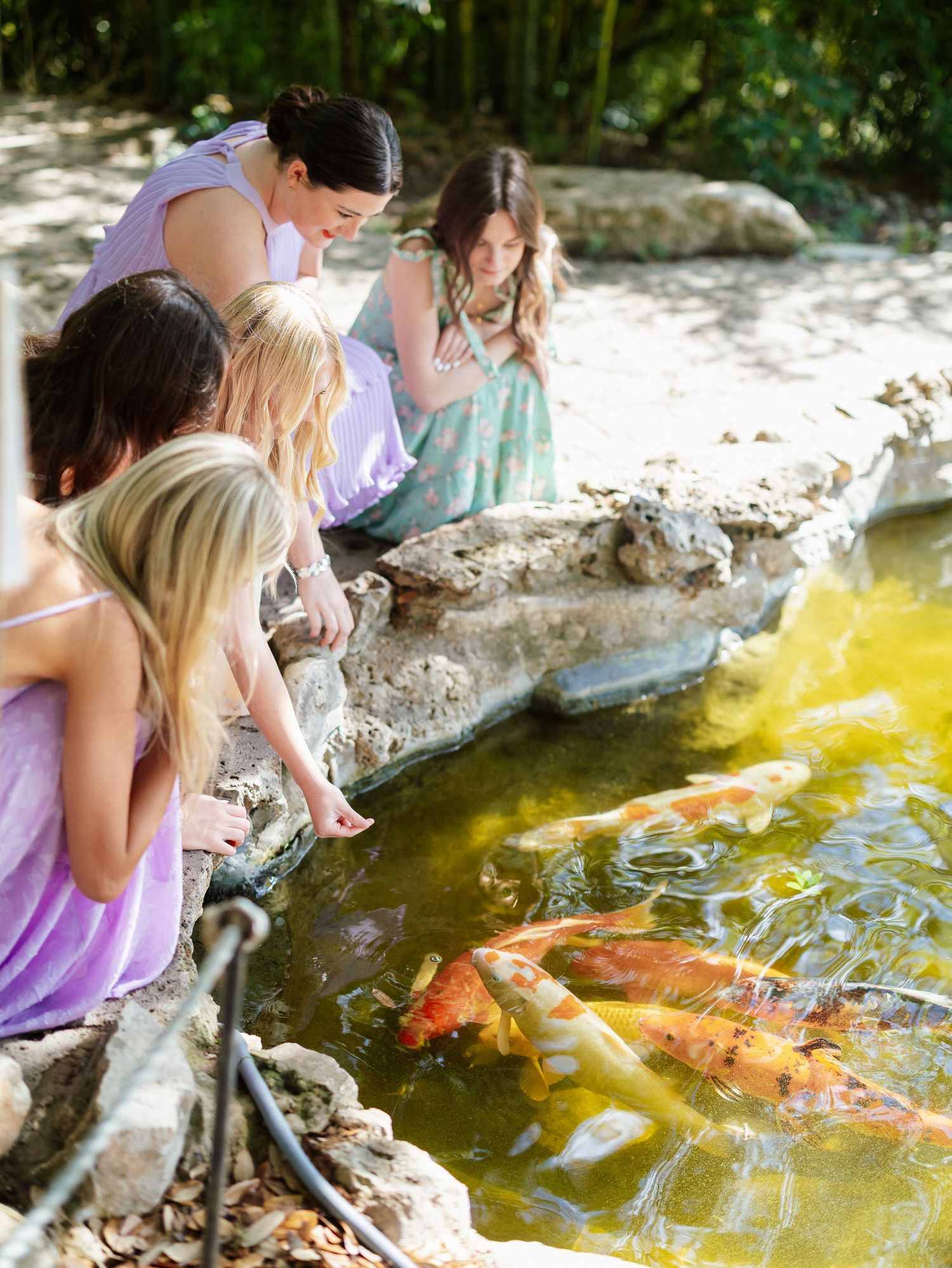 Girls feeding koi fish in a garden pond surrounded by rocks and lush greenery.