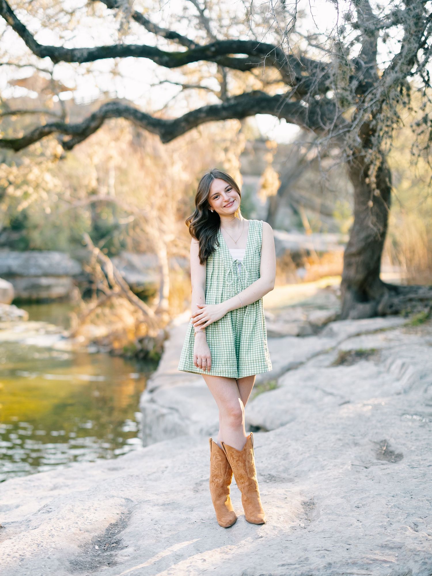 Young woman in green striped dress and cowboy boots posing on rocks near a creek with golden trees.