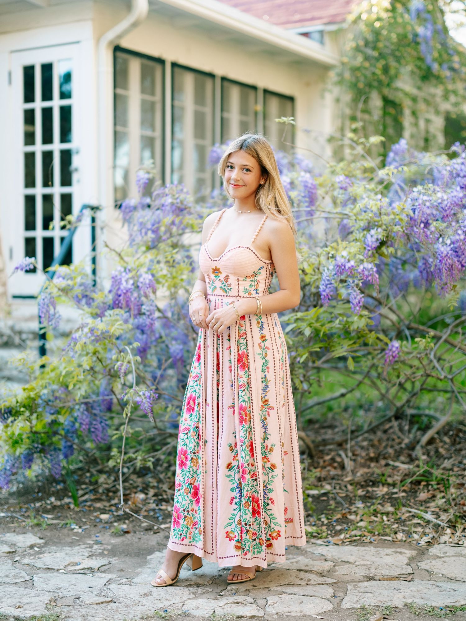 Woman in floral maxi dress posing in garden with purple wisteria blooms near a charming cottage.