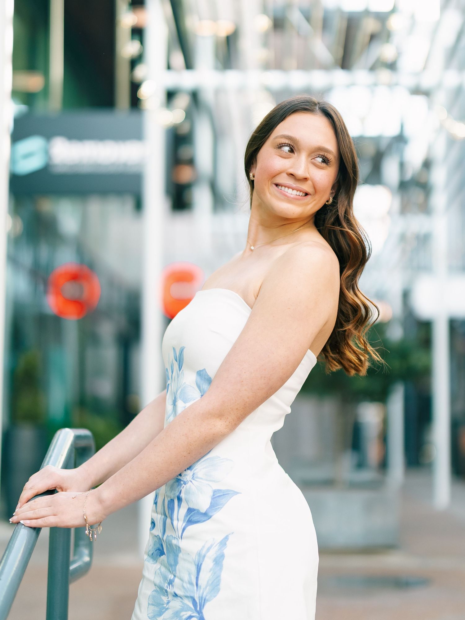 Smiling woman in strapless white floral dress posing outdoors near metal railing in urban setting.