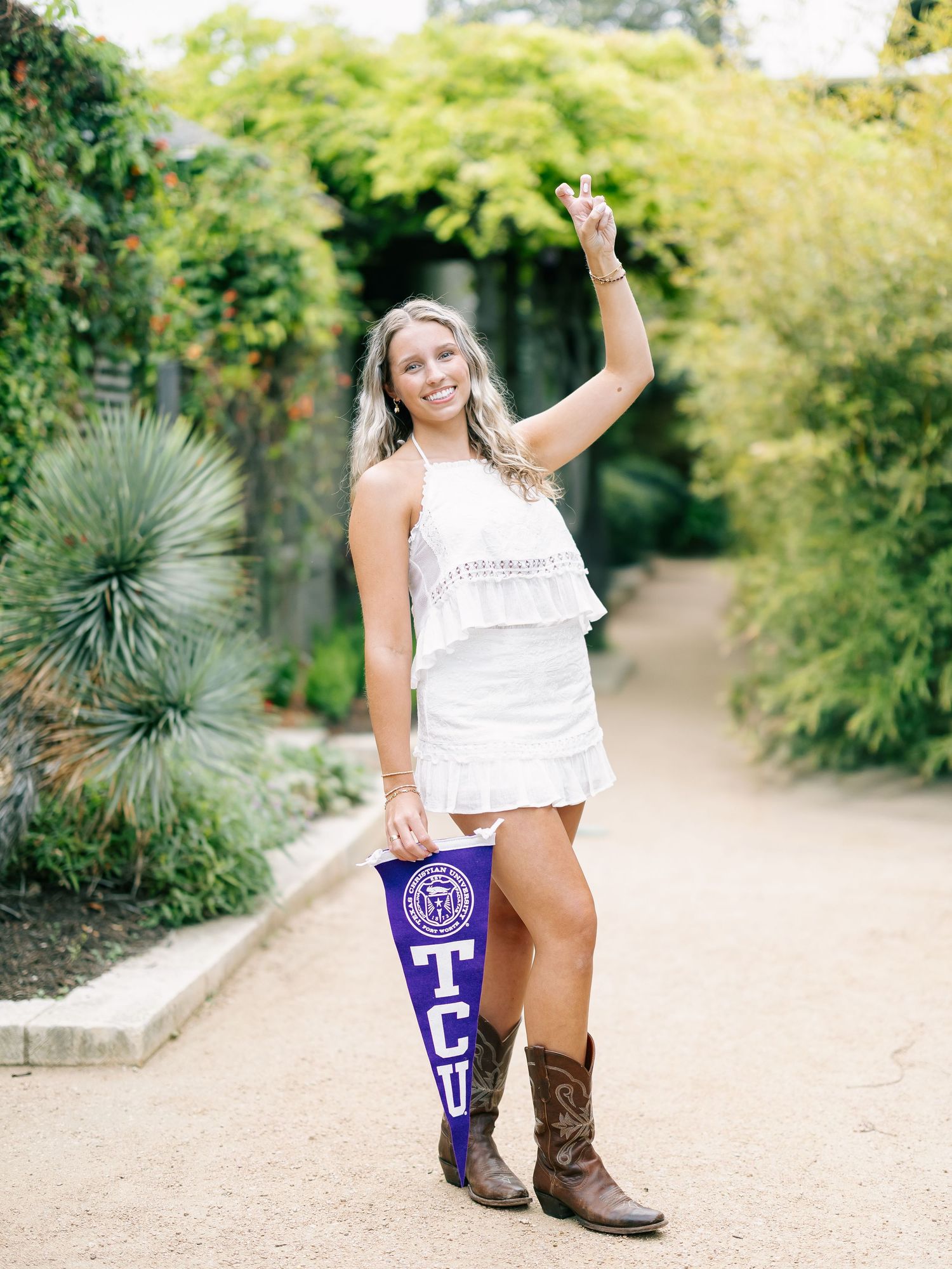 Smiling blonde woman in white dress holding TCU pennant and cowboy boots in a lush garden pathway.
