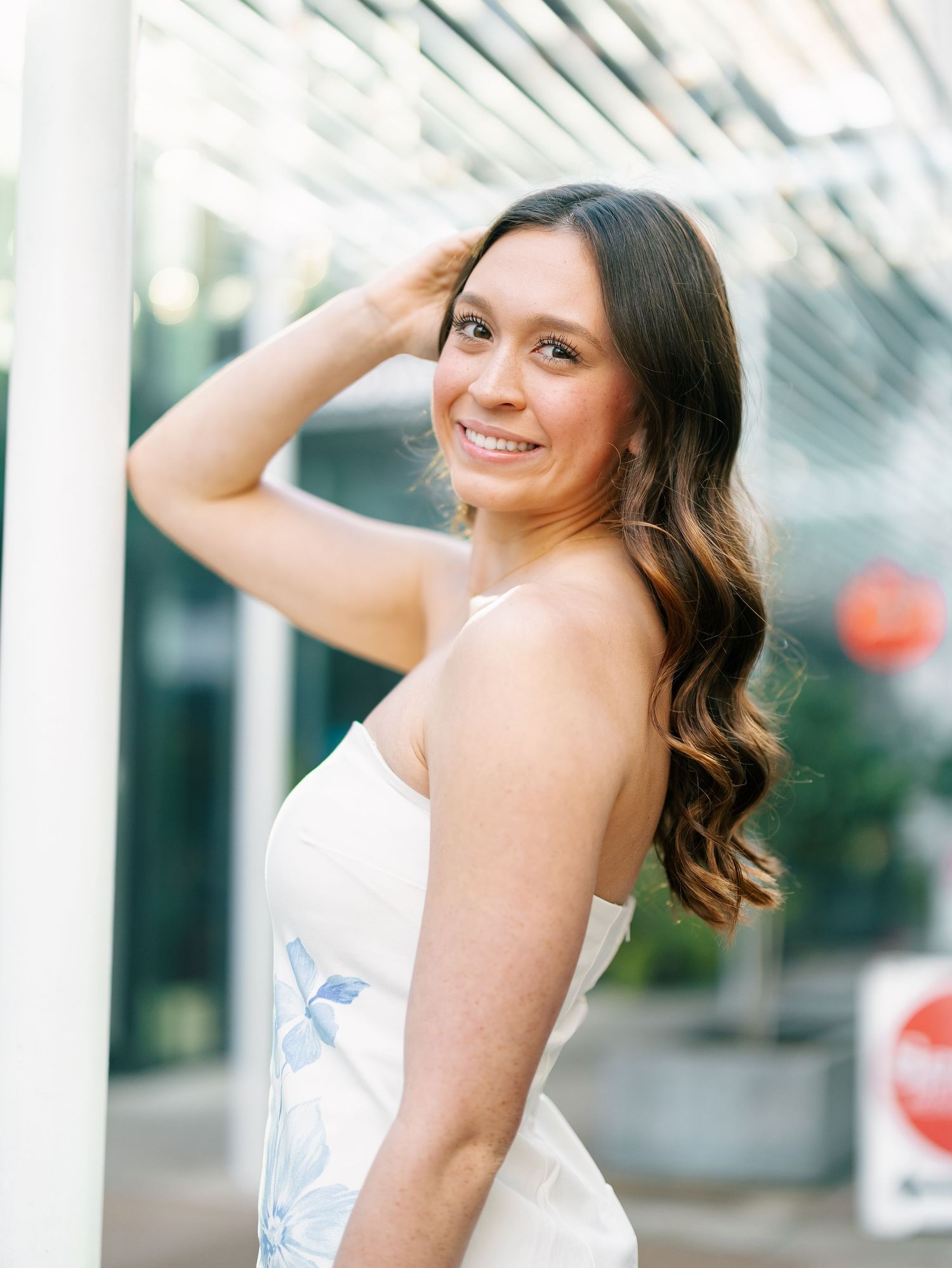 Brunette woman in white floral dress posing near pillar with hand in hair, smiling over shoulder.