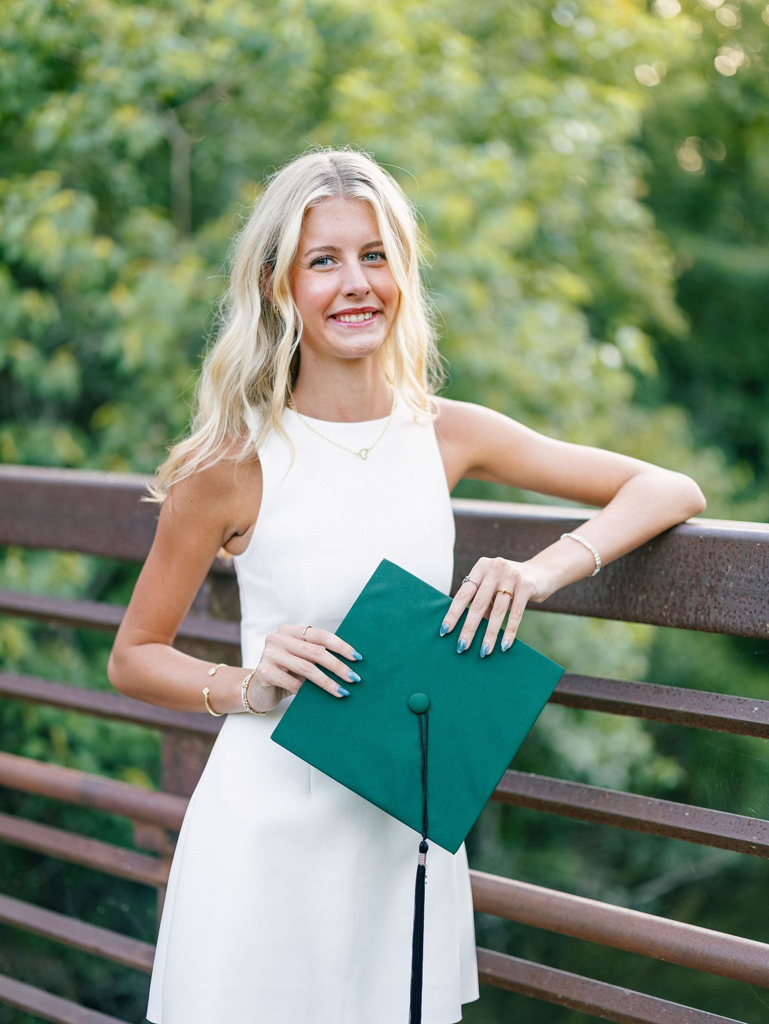 Smiling blonde woman in white dress holding a green graduation cap on a wooden bridge.