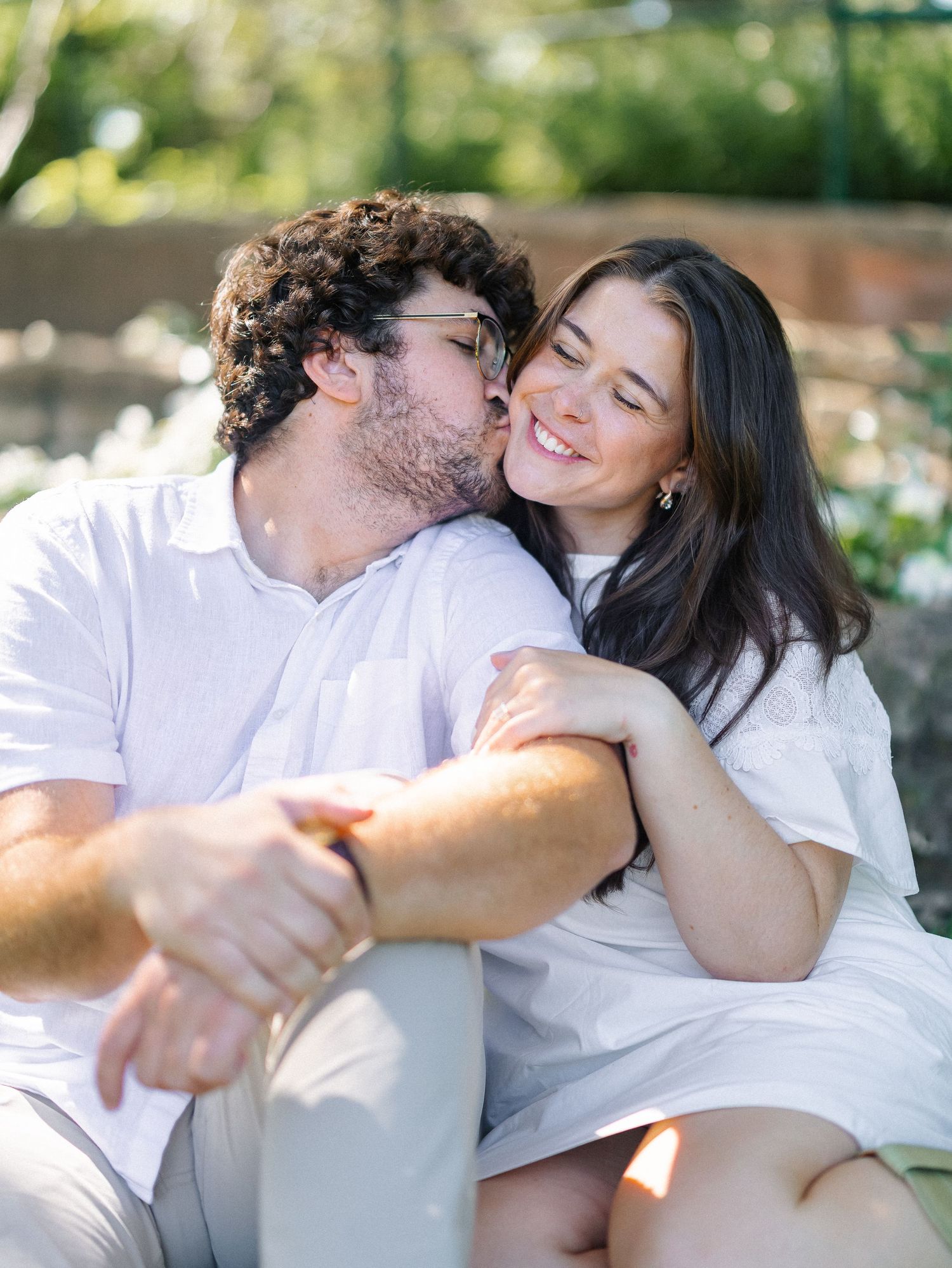A couple in white outfits share a tender moment outdoors, man kissing woman's cheek as she smiles warmly.