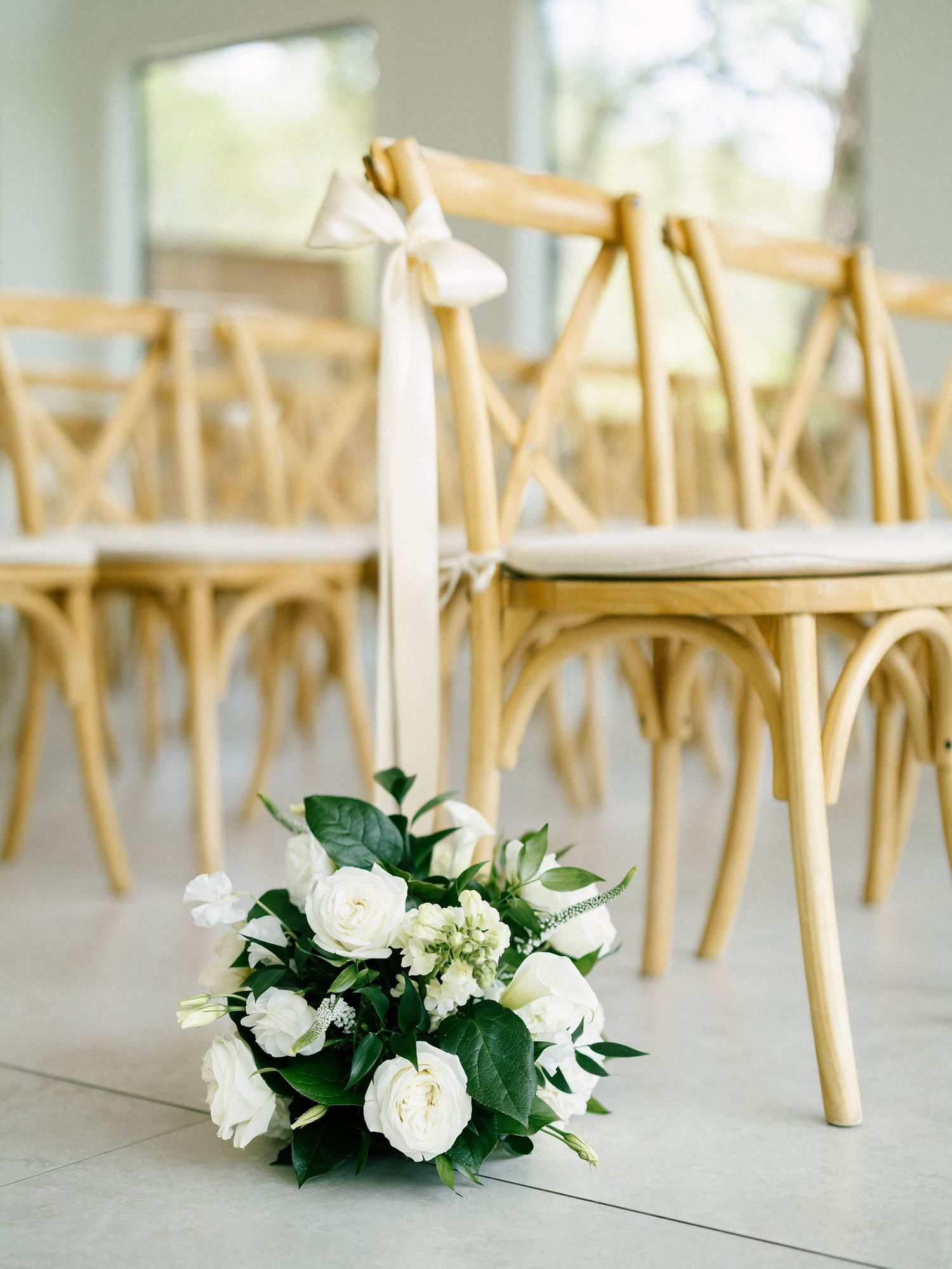 White rose bouquet with greenery on floor beside natural wood cross-back chairs at an elegant wedding ceremony.