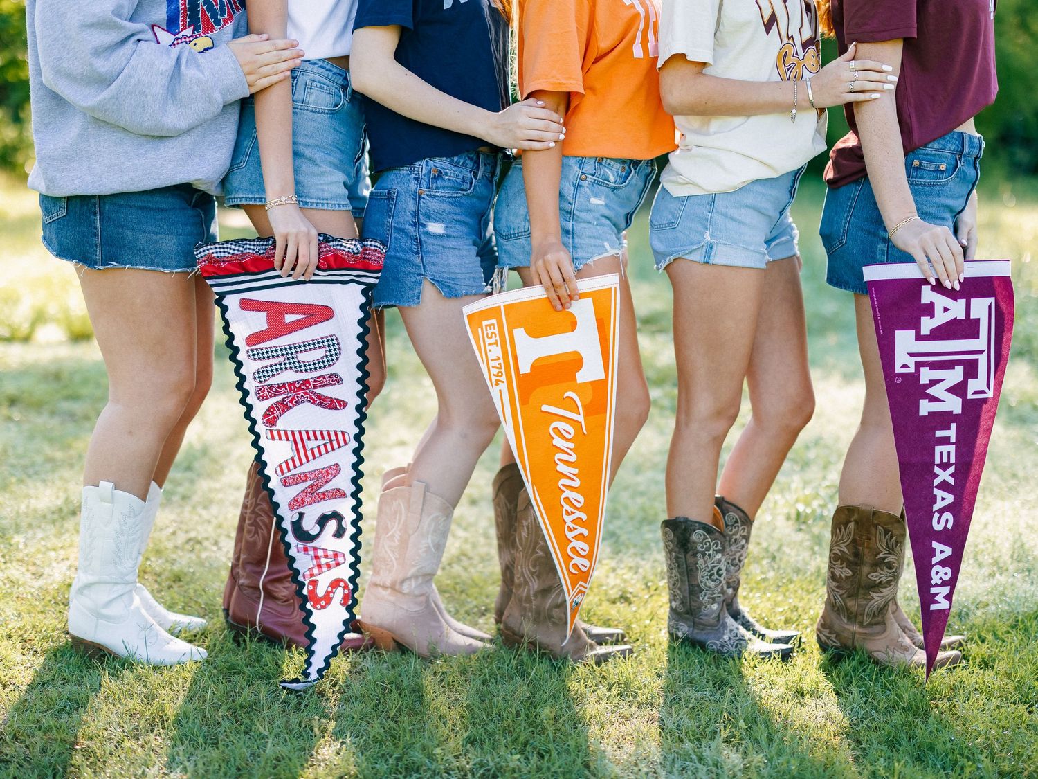 Group of girls in denim shorts and boots holding college pennants for Arkansas, Tennessee, and Texas A&M.