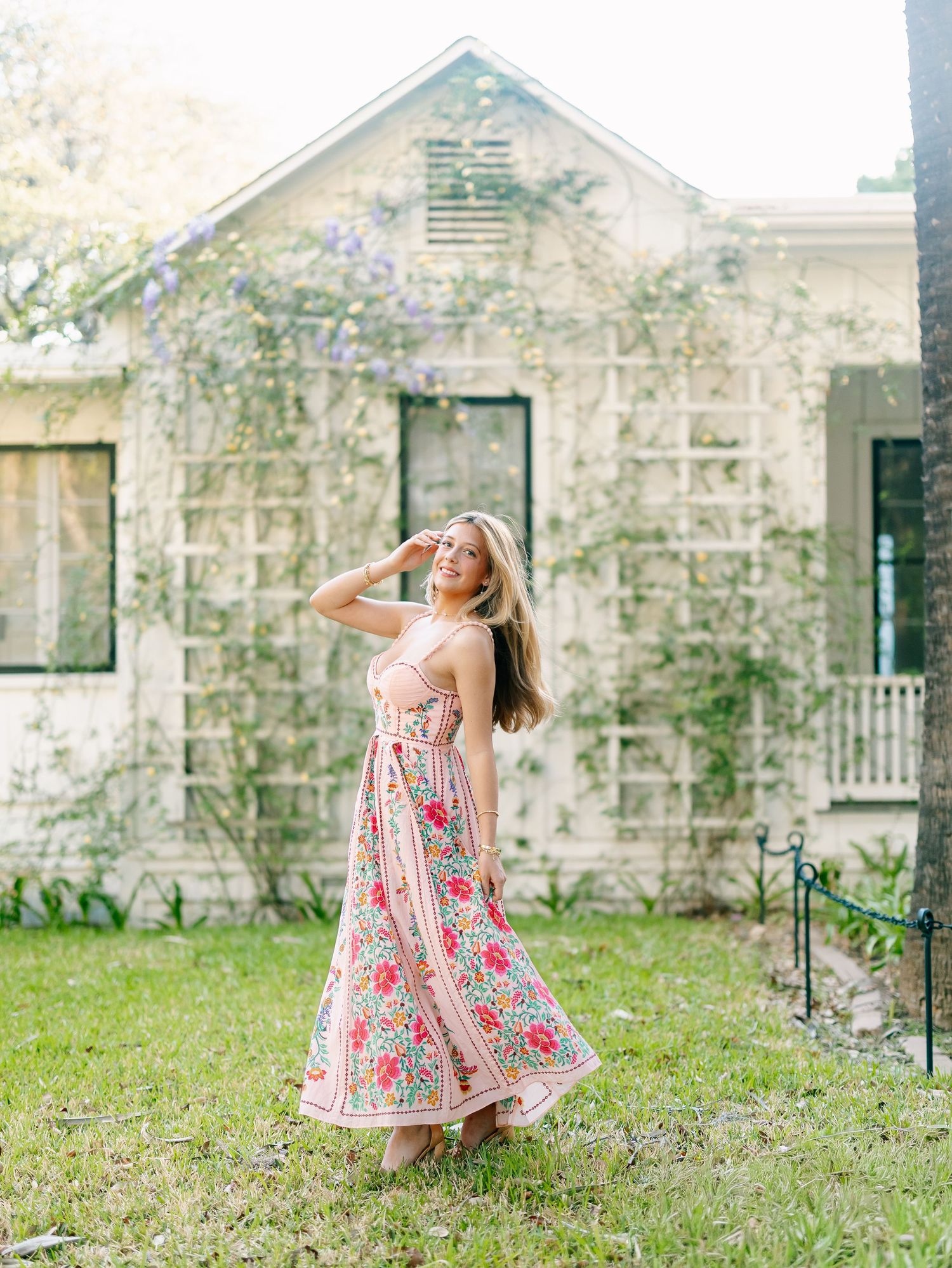 Young woman in floral maxi dress posing in front of a charming ivy-covered cottage with purple blooming vines.