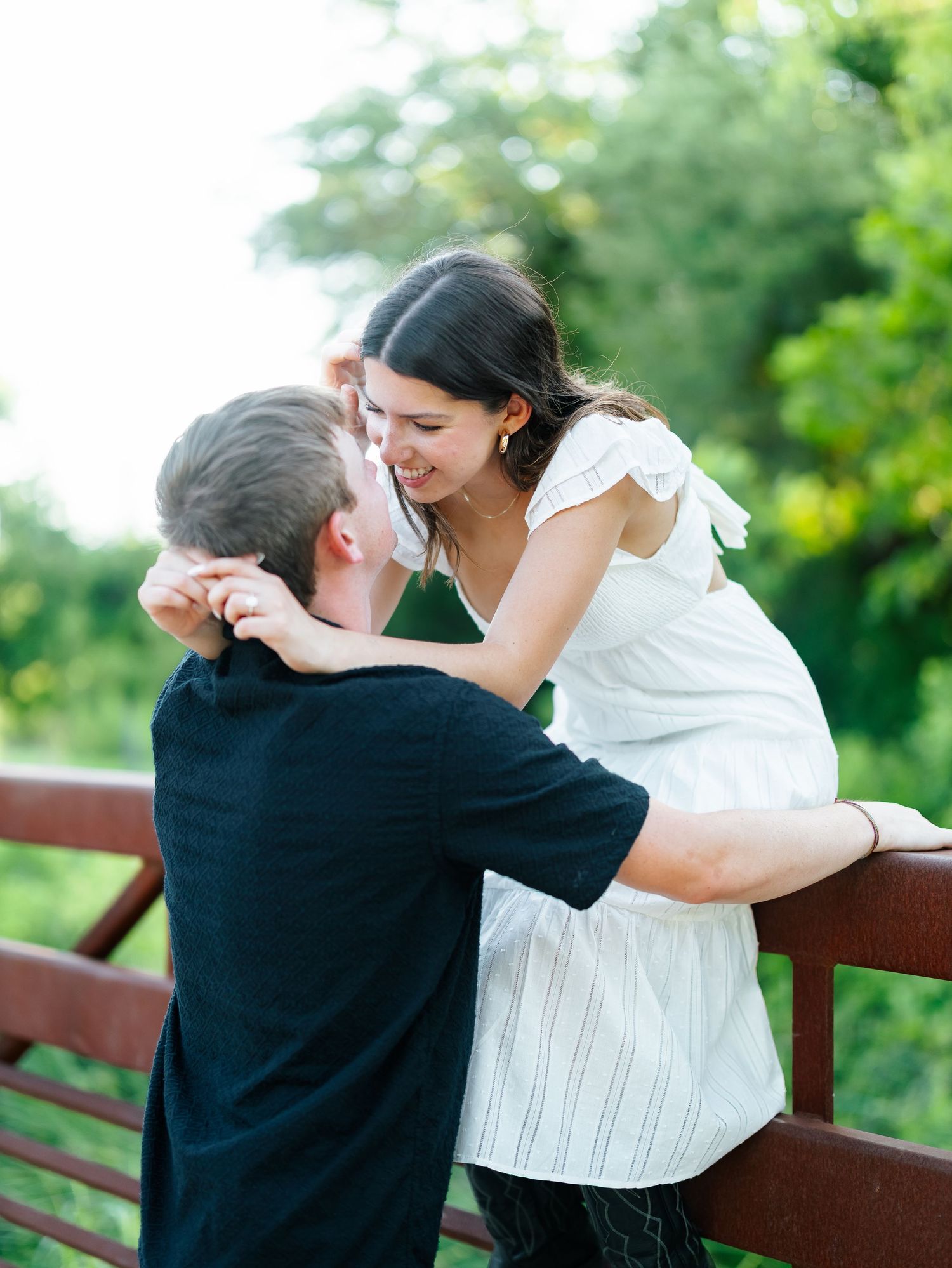 Couple smiling and leaning close together on a red wooden bridge surrounded by green trees.