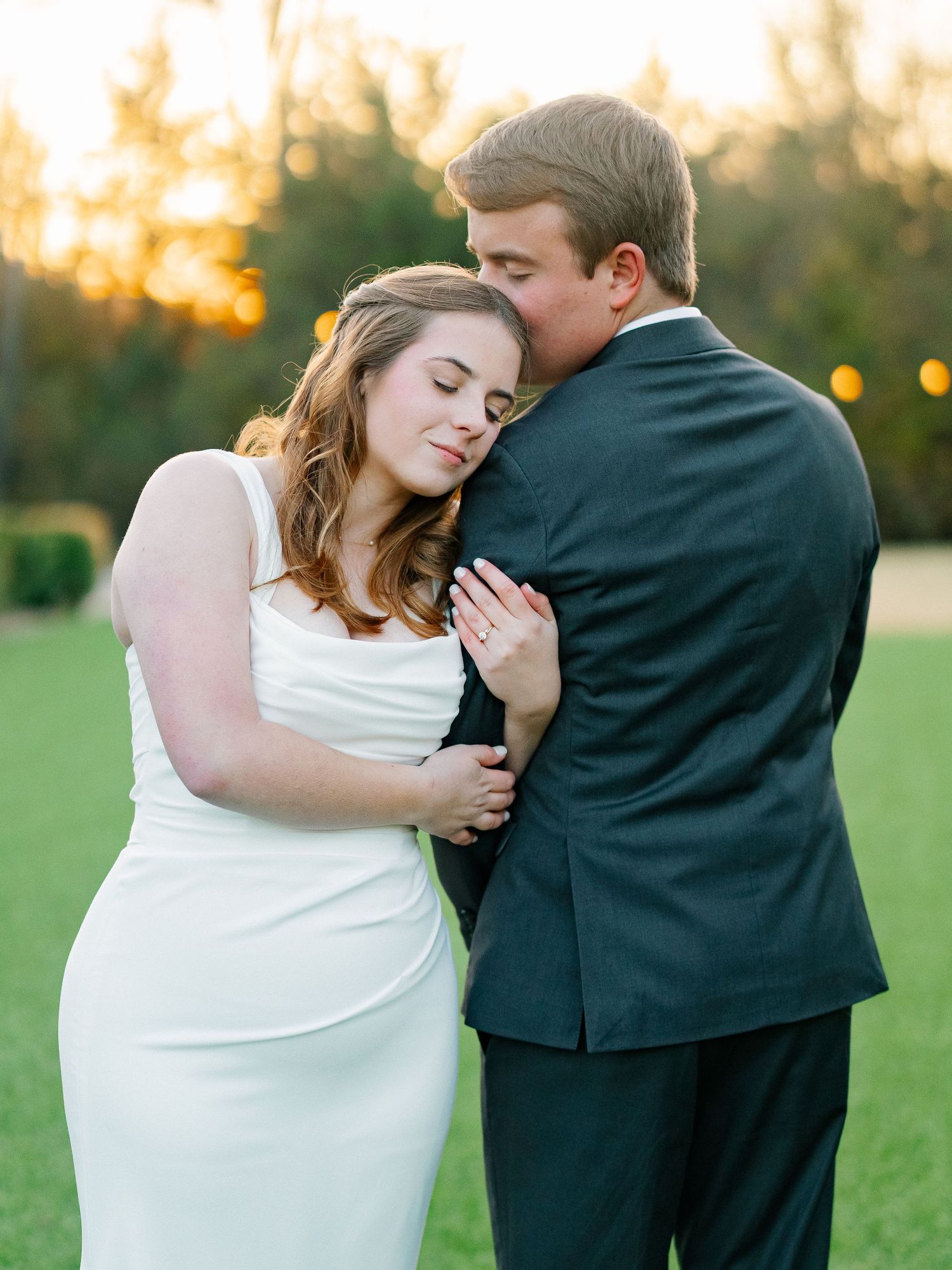 Bride in sleek white gown embracing groom in black suit during golden hour outdoor wedding portrait.