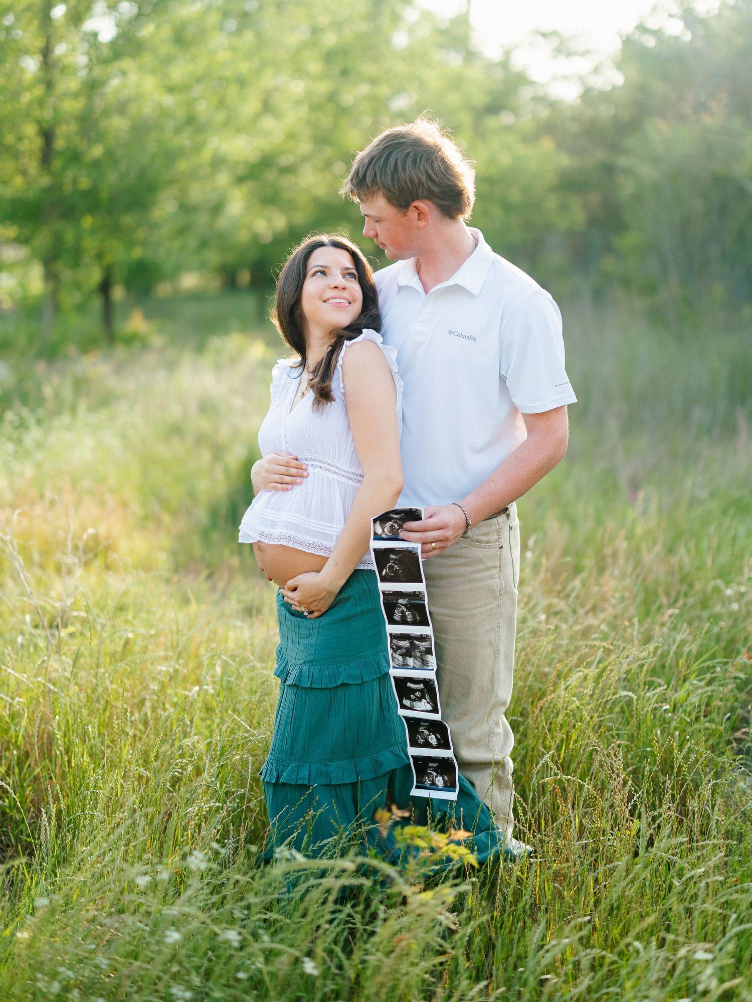 Expecting couple holding ultrasound photos in a sunny green meadow.